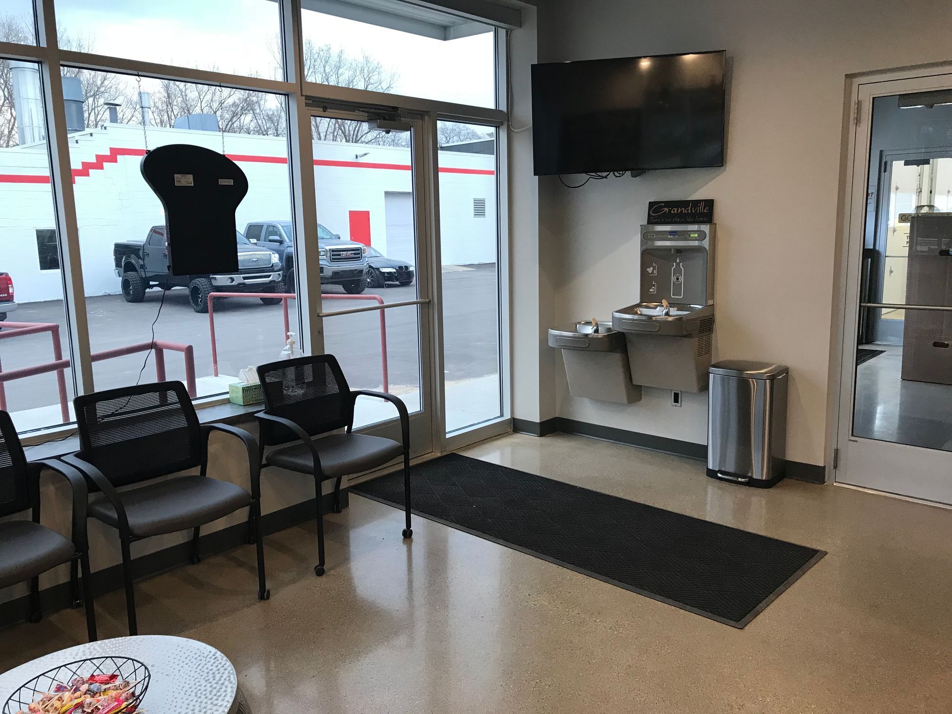 Waiting area with black chairs, front windows, wall TV, water dispenser, and a door mat on the floor