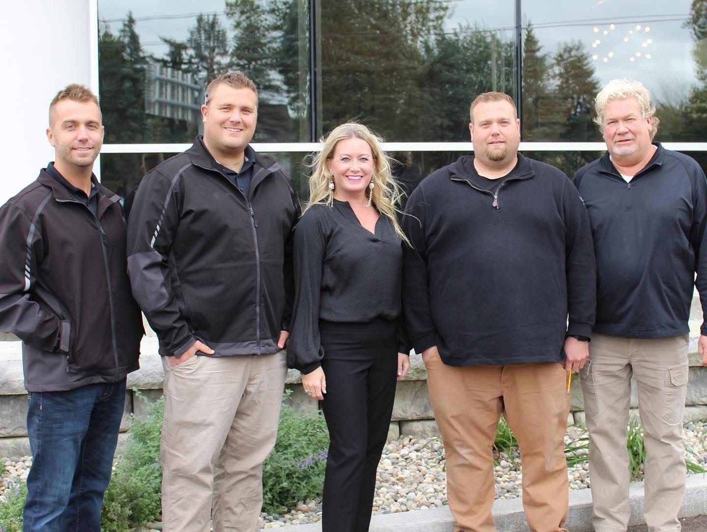 Five people standing outdoors in front of a glass building, wearing dark jackets and casual pants.