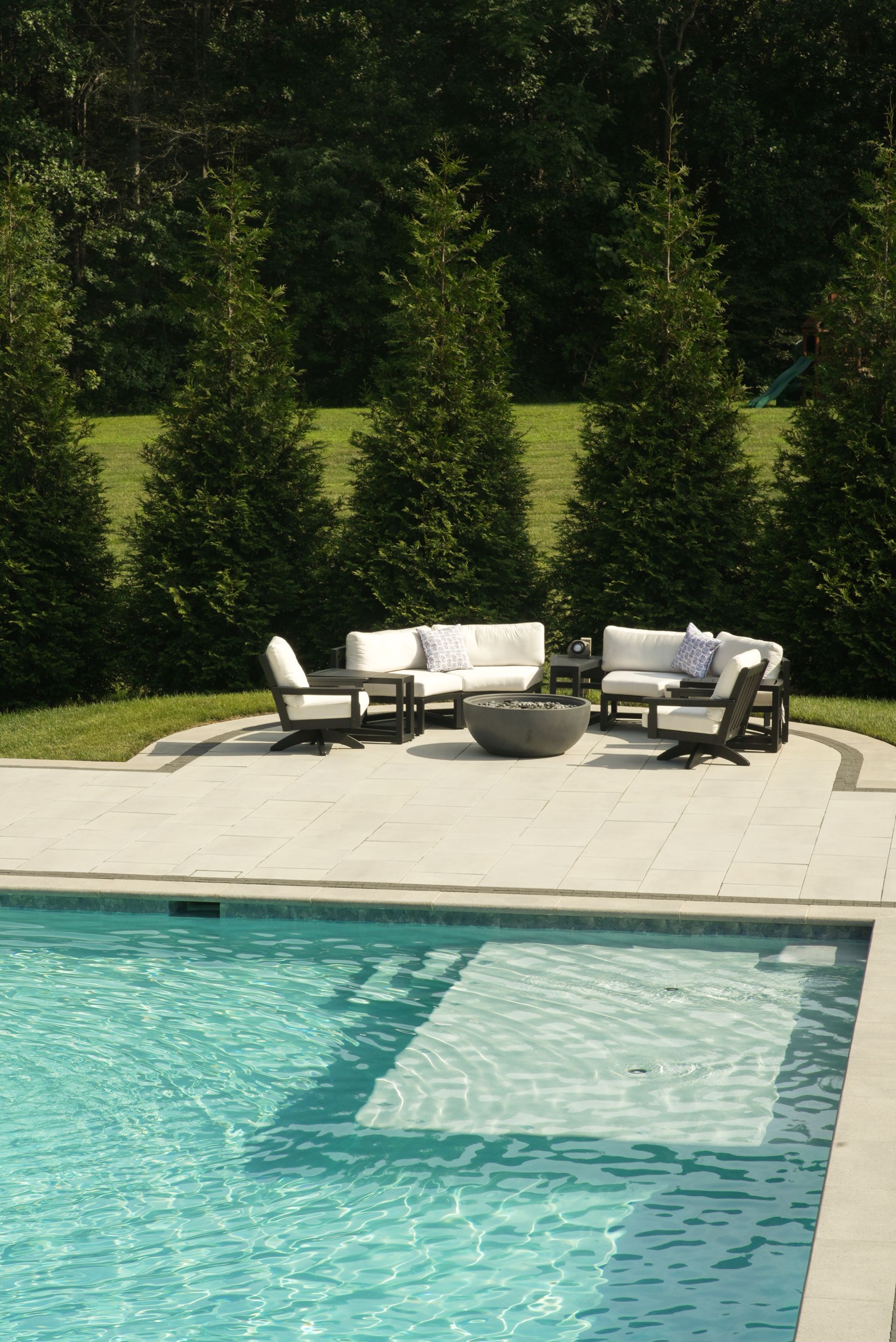 A pool area with white couches, a fire pit, and a row of green trees in the background.