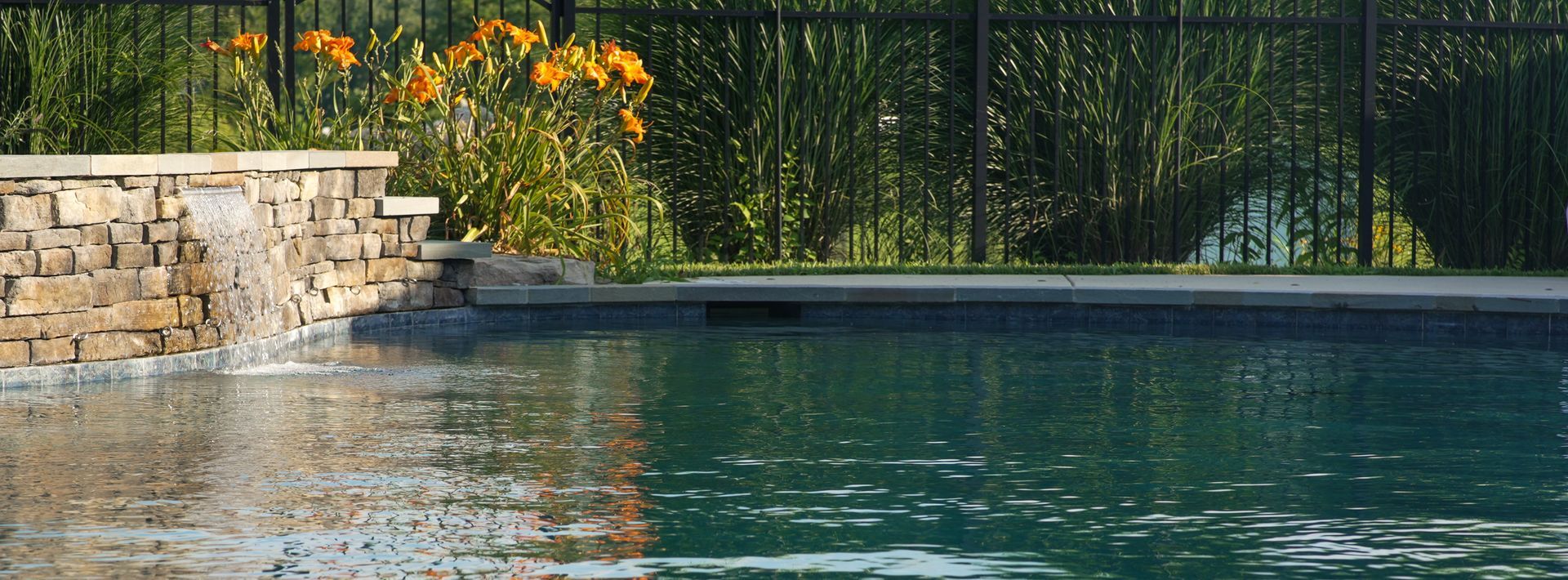 A stone wall with a waterfall cascades into a pool. Behind the pool is a green hedge and a black fence.