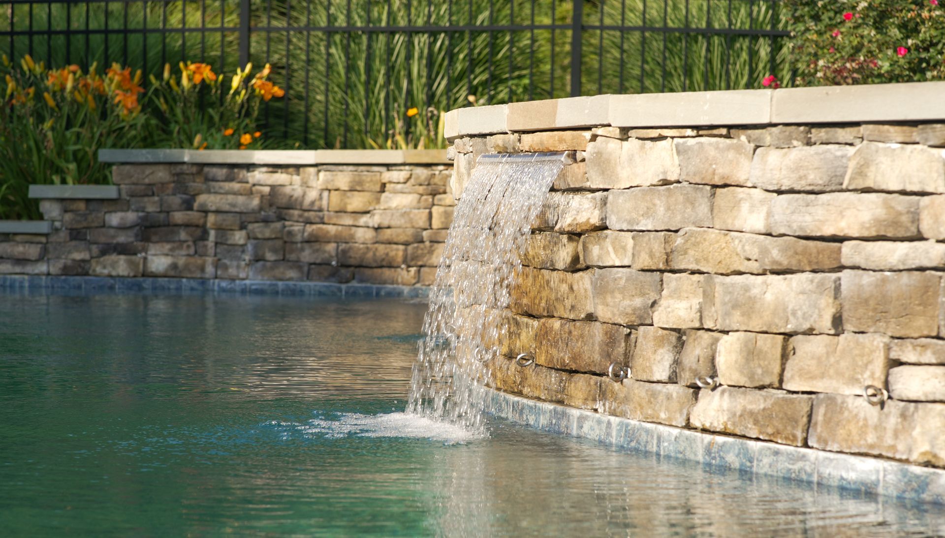 A stone wall with a waterfall cascading into a swimming pool. Yellow flowers and a black fence are in the background.