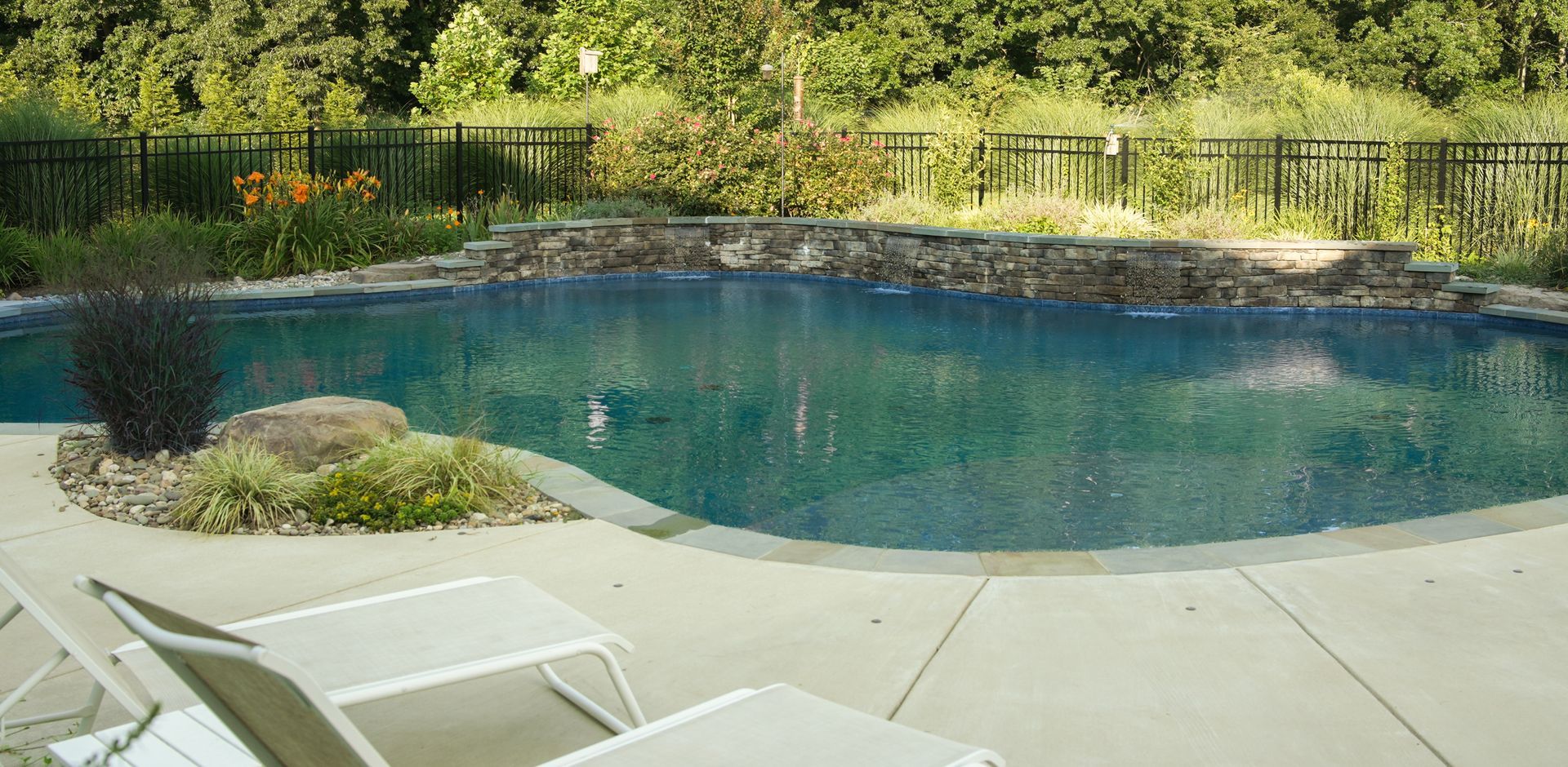 A swimming pool with a rock waterfall and surrounding foliage. Lounge chairs rest on the concrete patio.