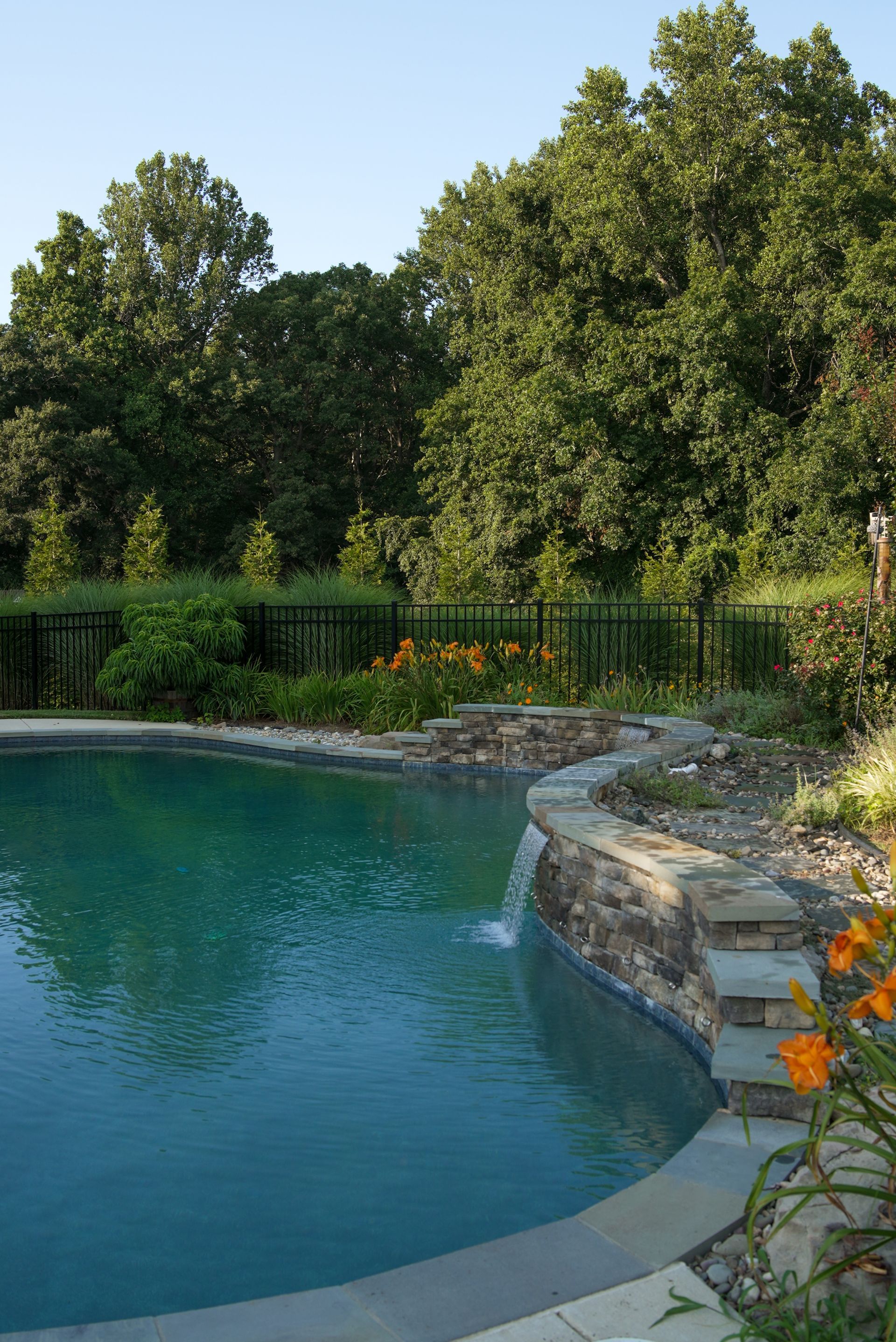 Pool with waterfall feature; surrounded by stonework and landscaping, backed by a fence and trees.