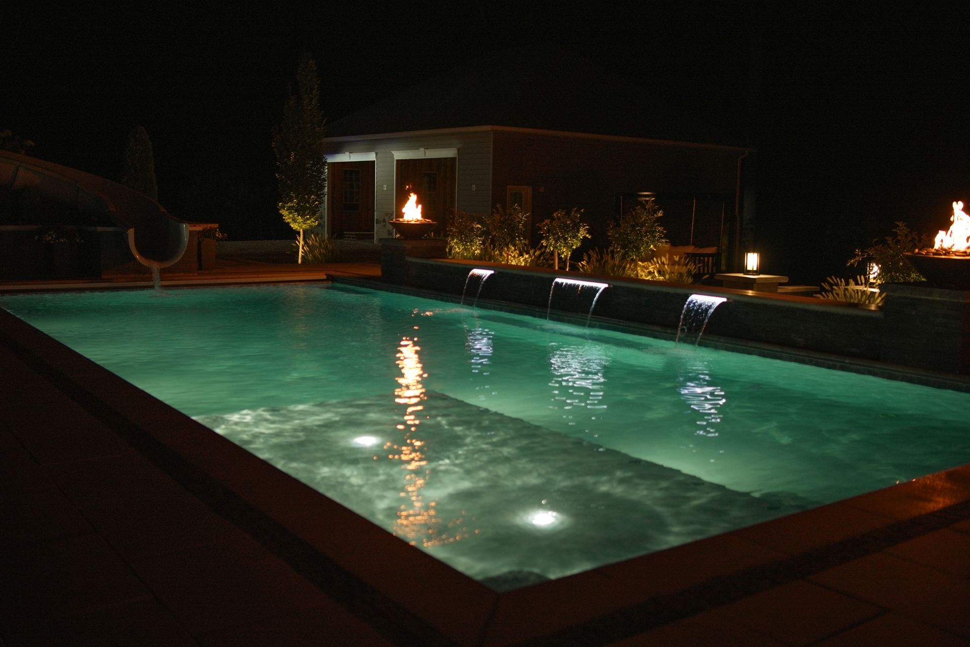 Nighttime view of a rectangular pool with cascading waterfalls and underwater lights, with a small structure and fire features in the background.