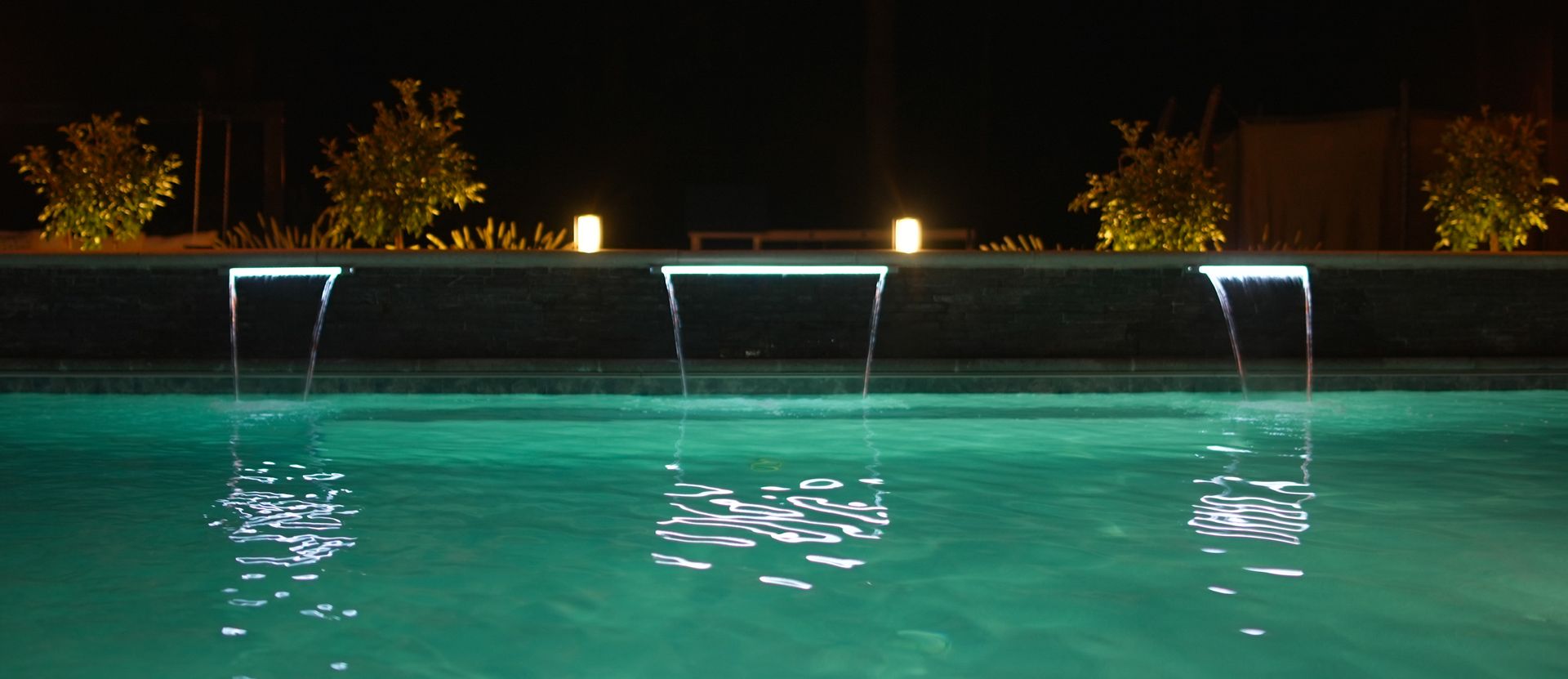A pool with illuminated water features at night. Water flows from a ledge into the turquoise water below.