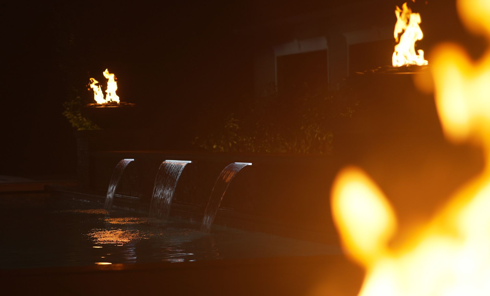 Waterfalls cascade into a dark pool, with flames atop a structure in the background. A large fire glows in the foreground, creating warm hues.