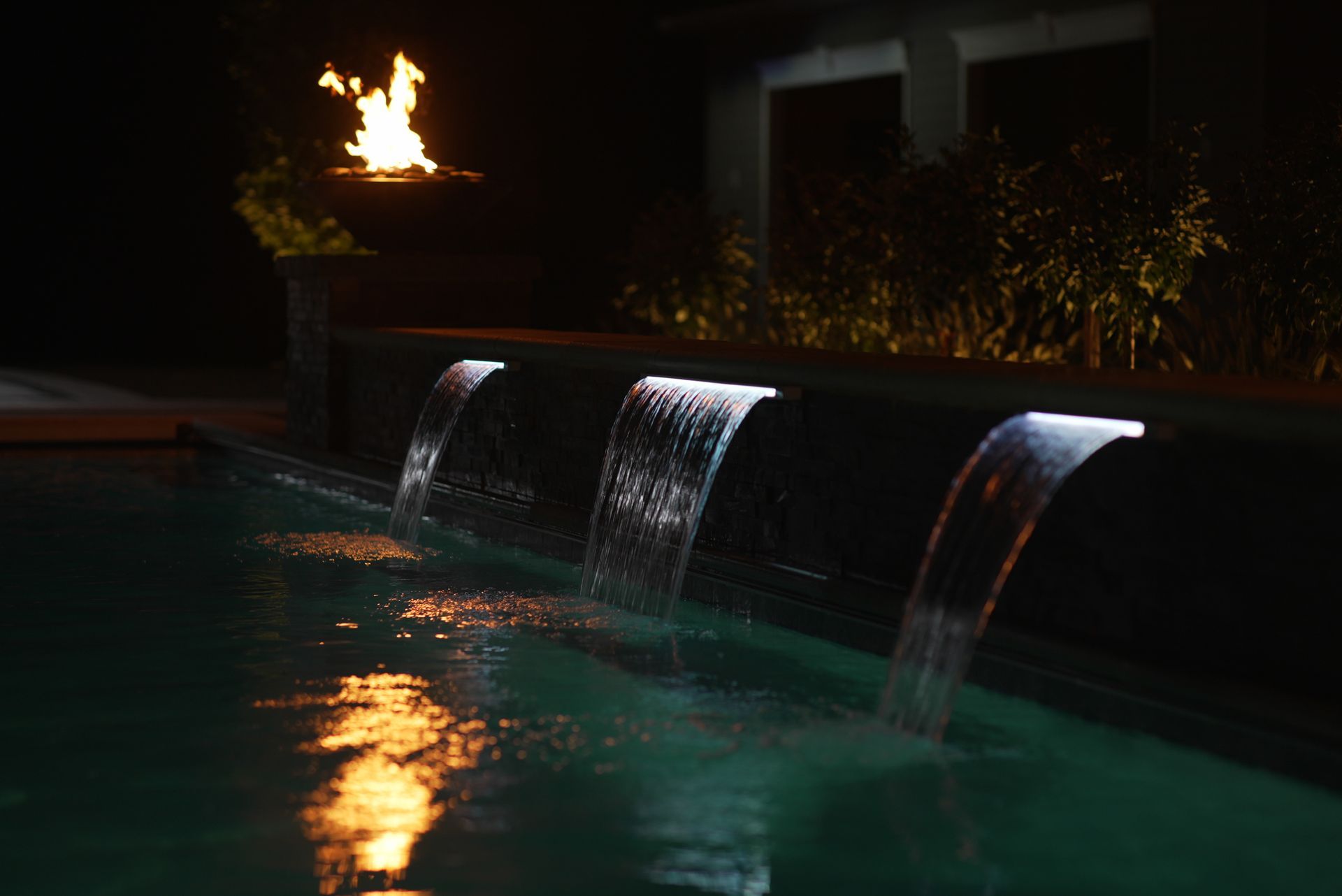 Pool with illuminated waterfalls at night, fire pit visible in the background. Teal water reflecting firelight and lighting.