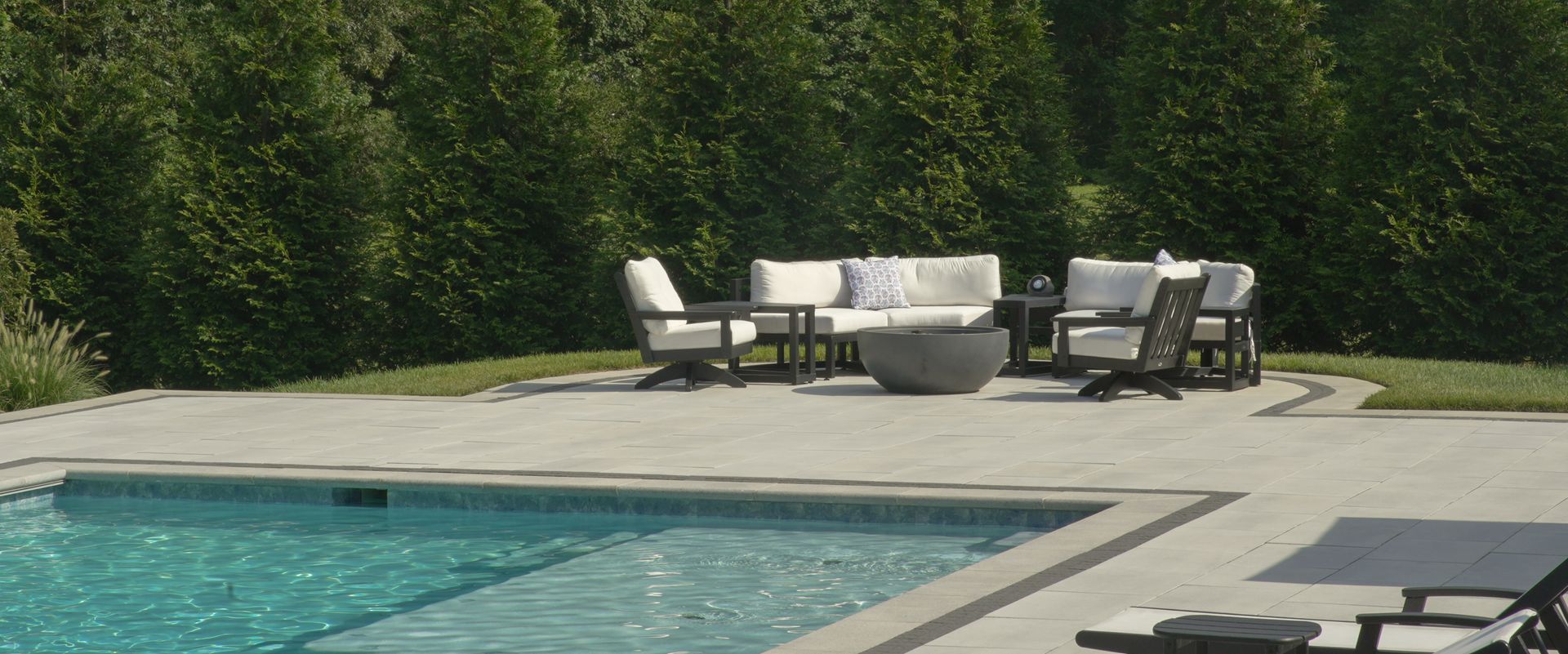 A backyard with a swimming pool and patio furniture. White couches and chairs are arranged around a fire pit. Green trees fill the background.
