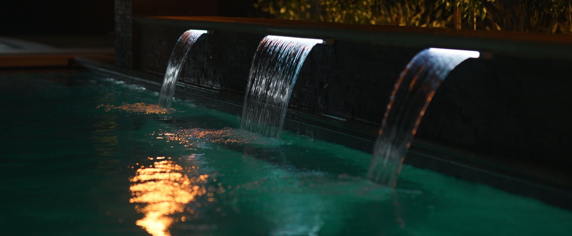 A nighttime pool with illuminated waterfalls cascading into the turquoise water. The scene is dimly lit with reflections.