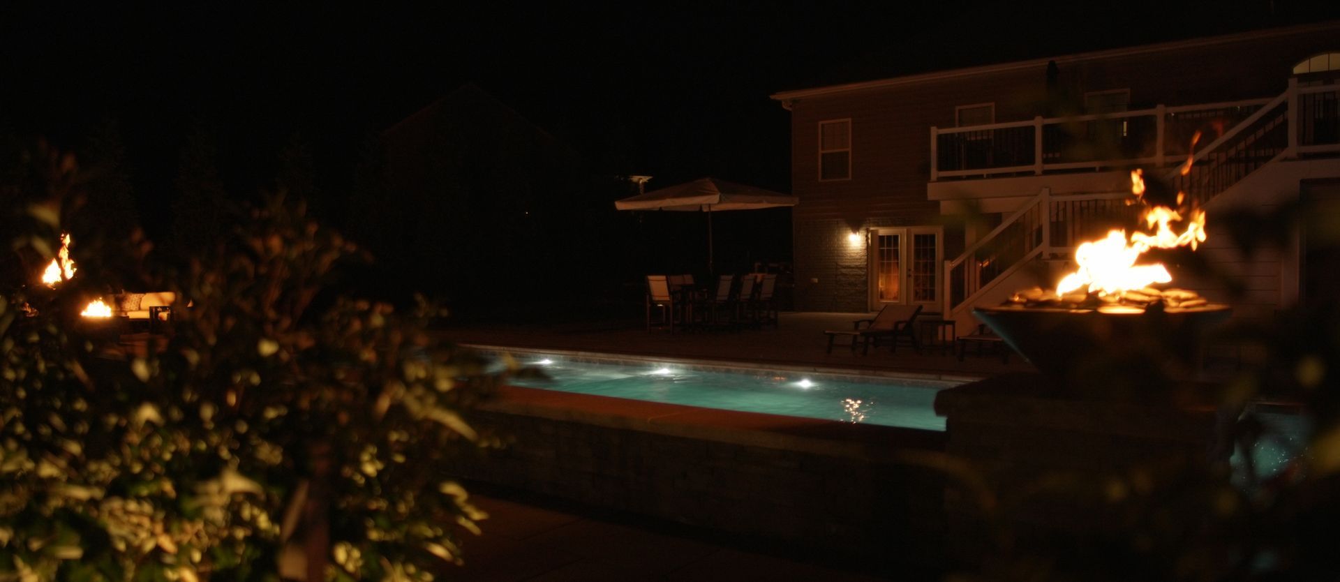 A nighttime scene of a pool and a house with a fire pit. The pool has lights in it, and the house has lit windows.