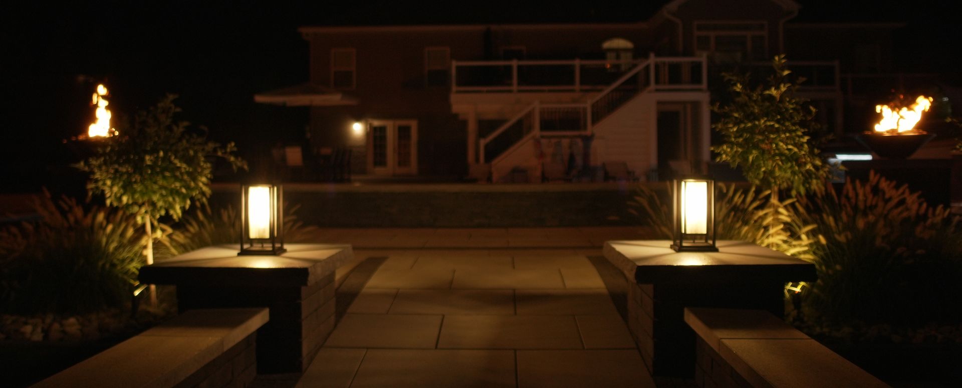 Nighttime view of an illuminated walkway leading to a house. Modern lanterns and torches provide light and a warm atmosphere.