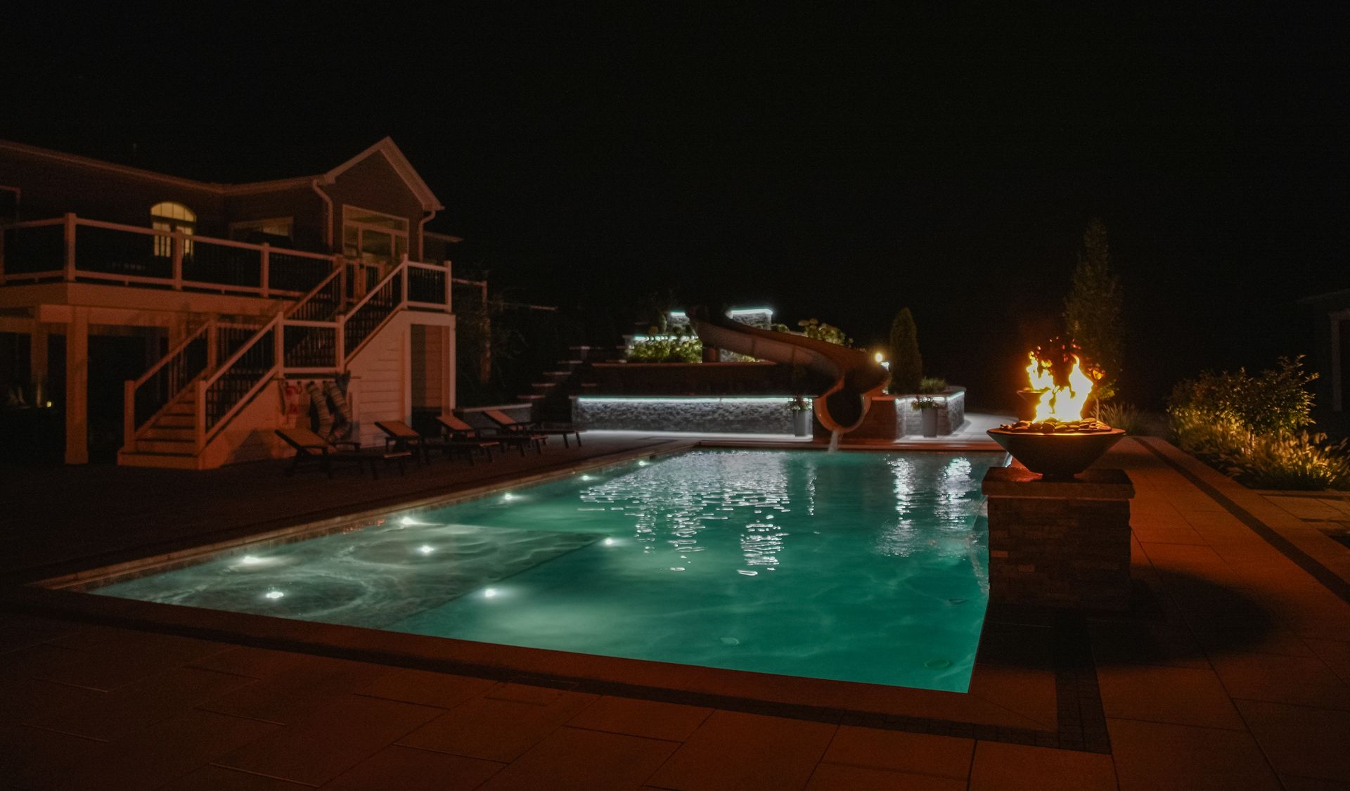 A nighttime view of a pool illuminated by lights, with a lit fire pit on the right, and a house in the background.