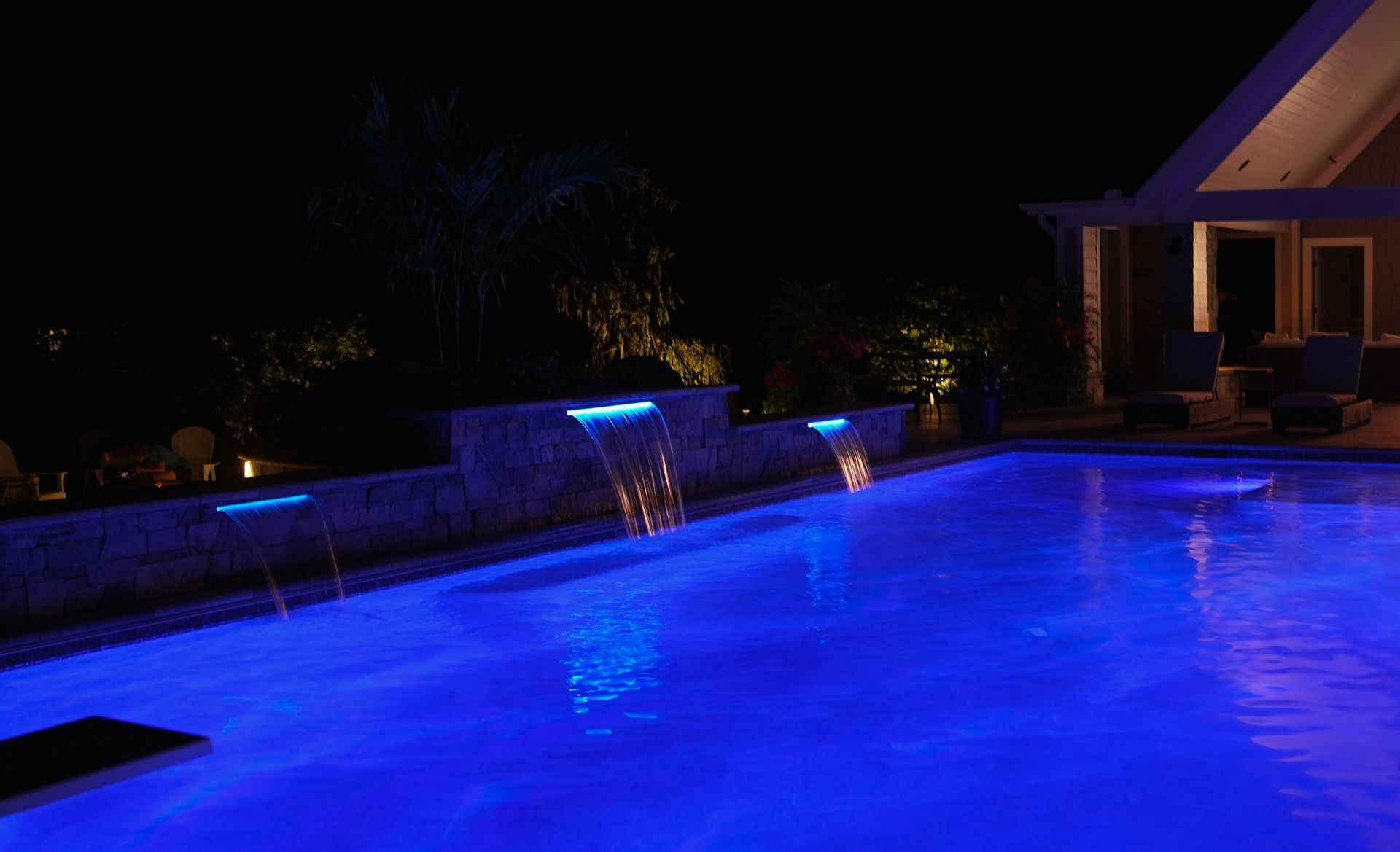 A nighttime view of a pool illuminated with blue lights, featuring waterfalls and the outline of a house.