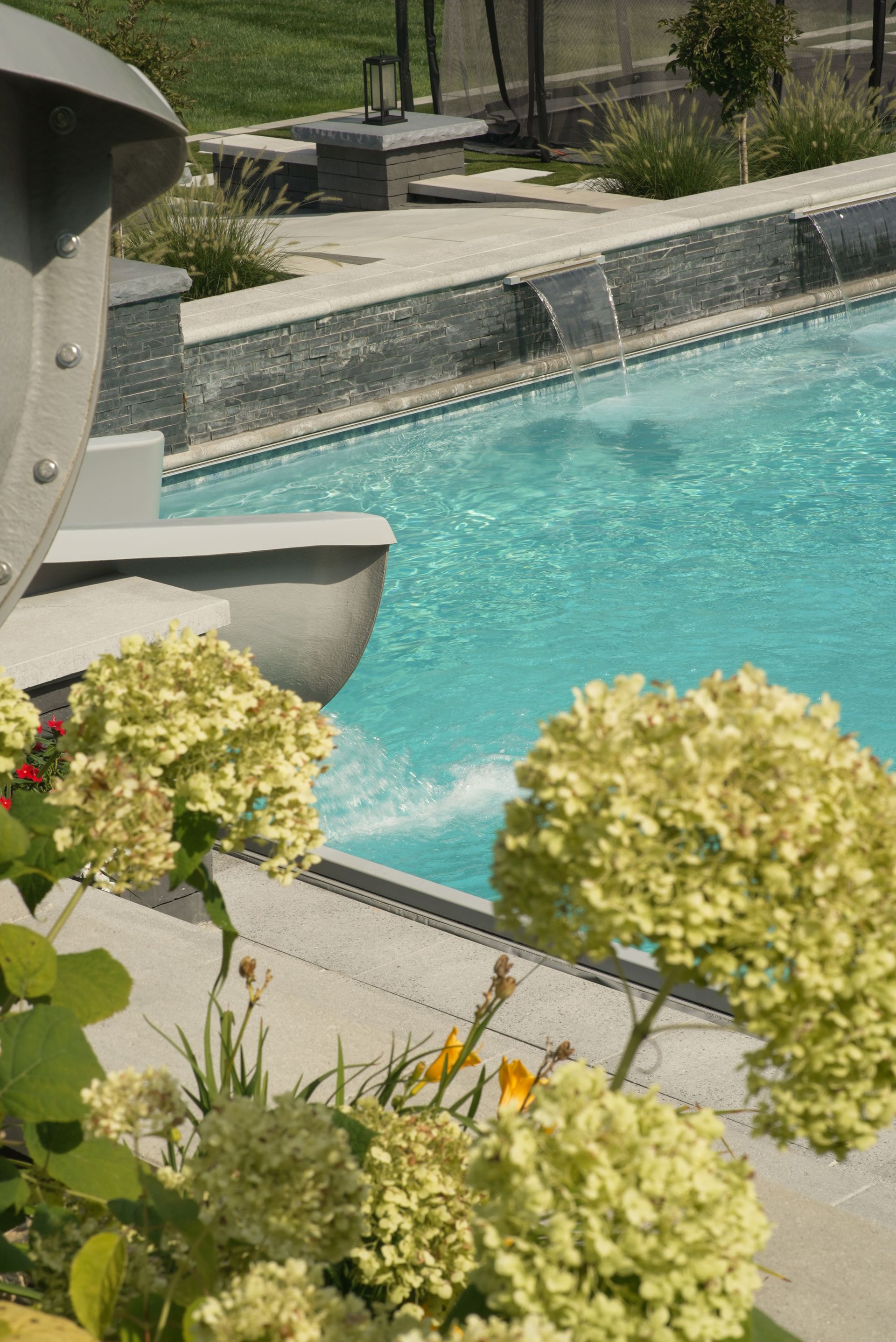 A pool with turquoise water and gray stone trim, surrounded by light green hydrangea flowers and greenery.