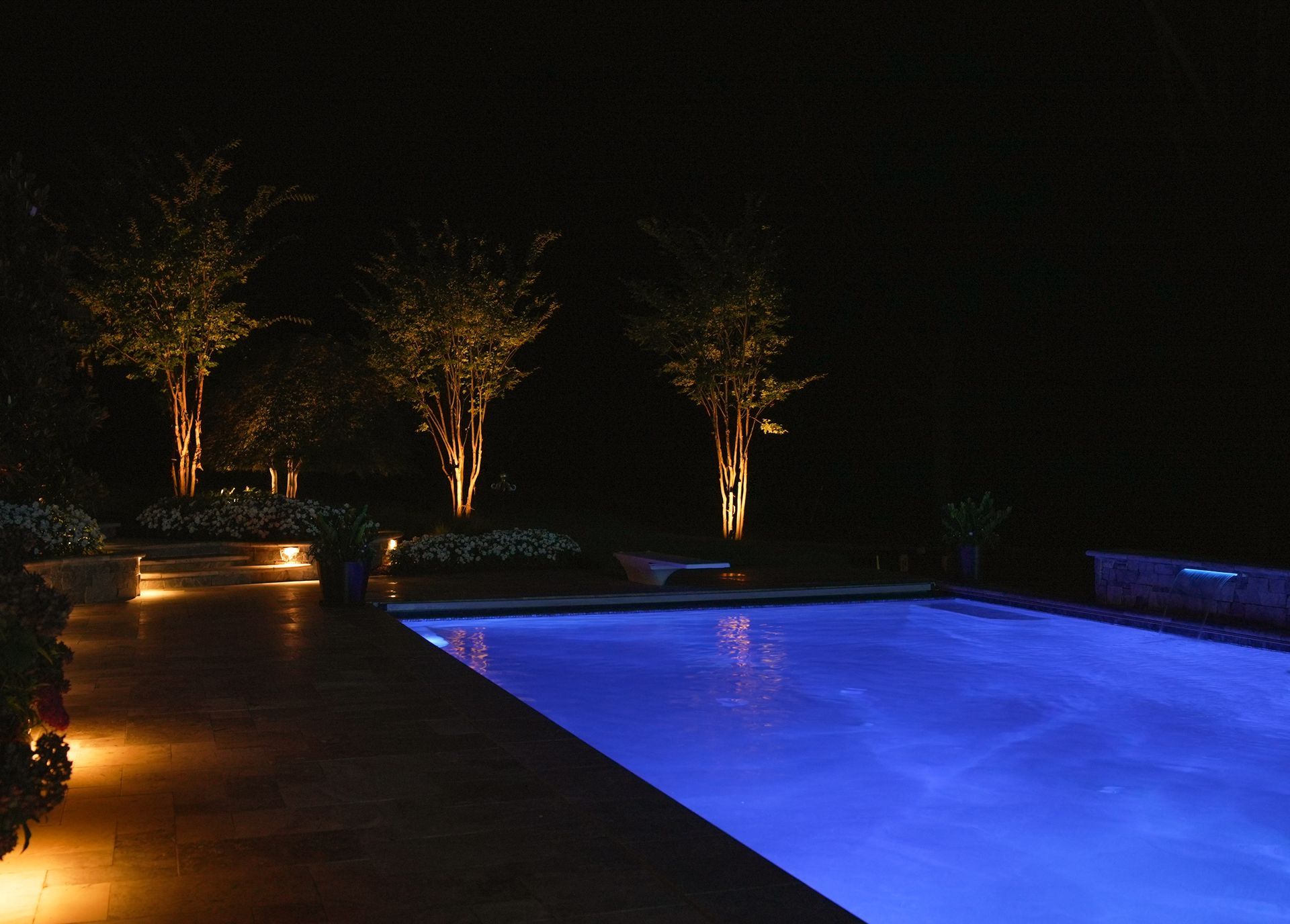Pool area at night lit with blue pool lights and spotlights illuminating trees and landscaping.