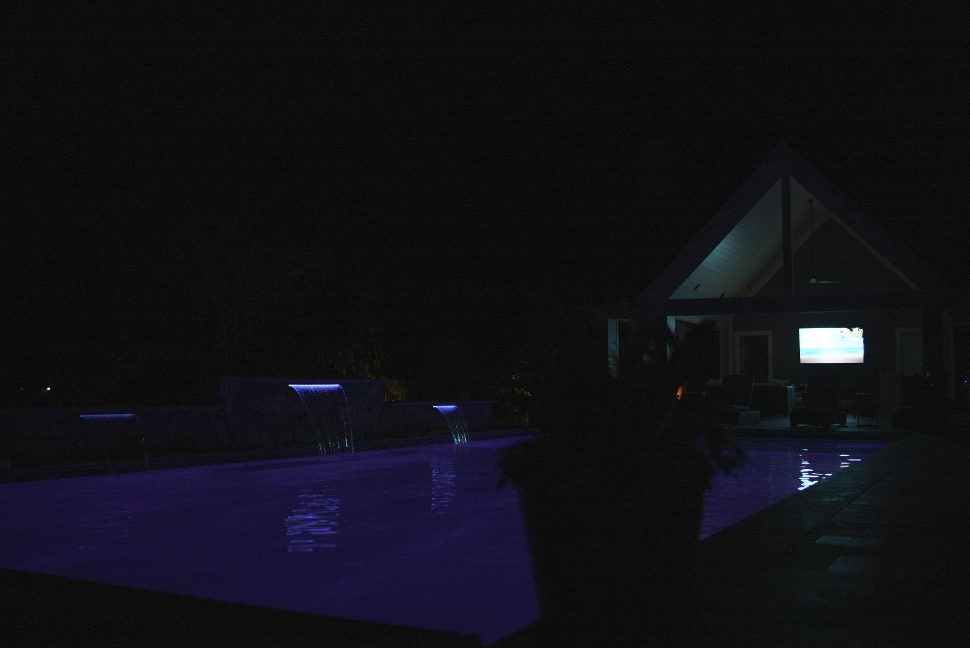 A nighttime view of a pool illuminated by purple lights, with a house in the background also lit up.