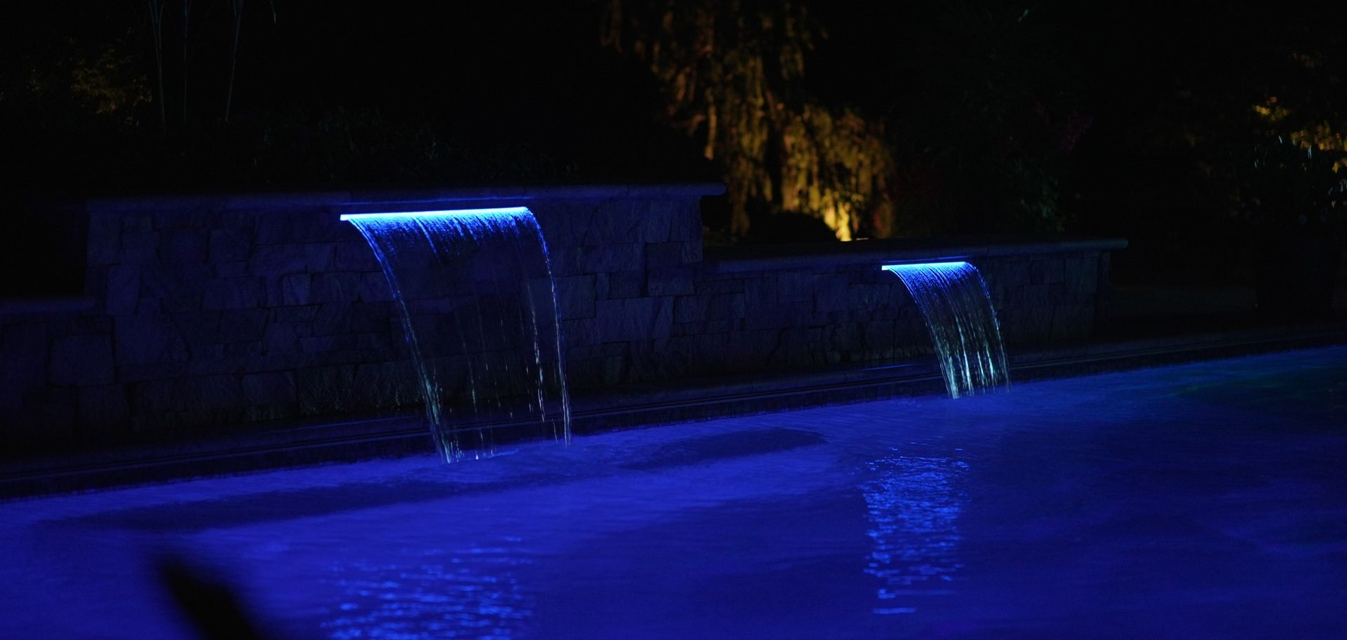 A nighttime pool with illuminated blue waterfalls cascading into the water.