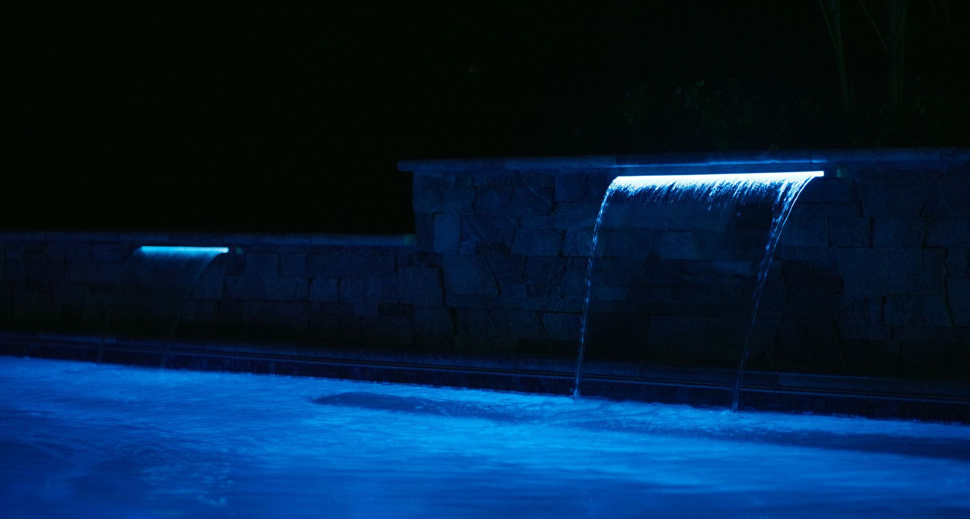 Blue-lit waterfall cascading into a pool at night. Dark surroundings with faint light highlighting the pool.