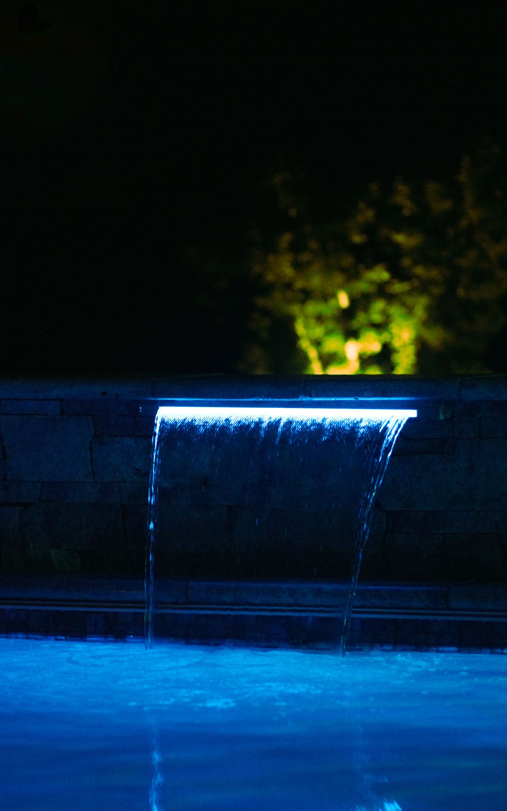 A small waterfall illuminated with blue light cascades into a pool at night. Dark background with a hint of green foliage.