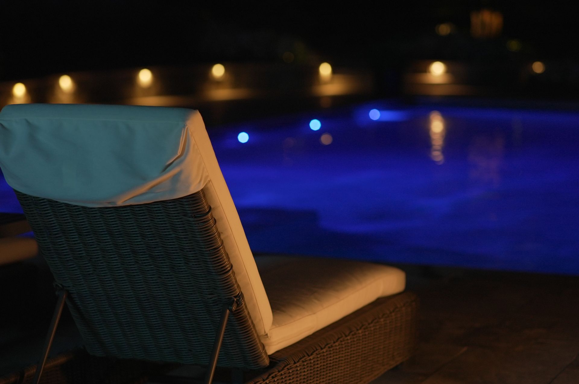 Lounge chair by a pool at night. The water is blue, and lights are reflected in the water.