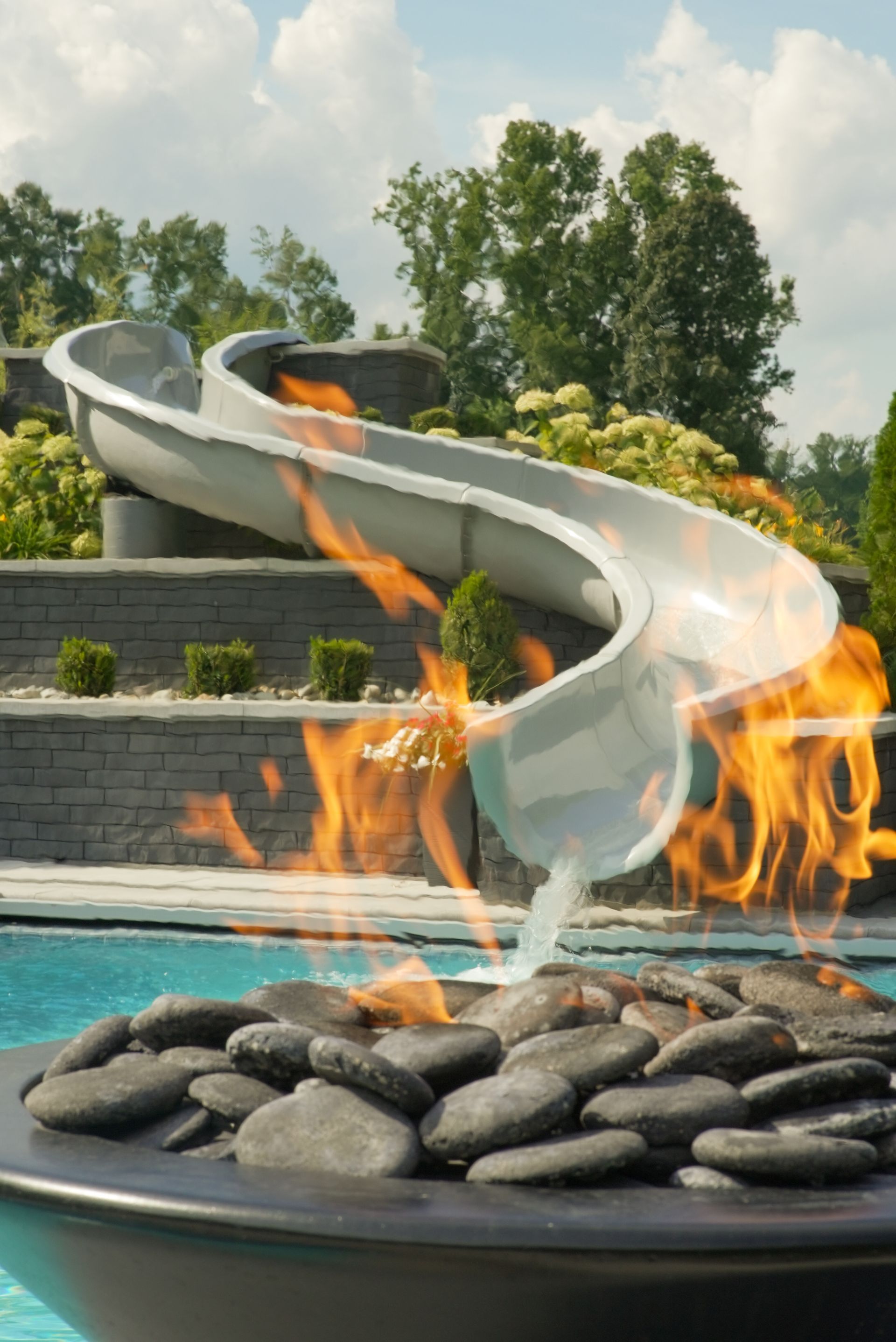 A swimming pool with a slide that leads into the water over a fire pit, surrounded by rocks and lush landscaping.