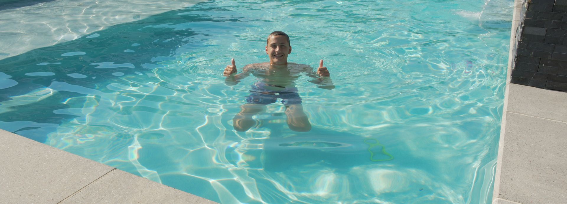 A young boy in a swimming pool gives two thumbs up. He is wearing blue swim trunks.