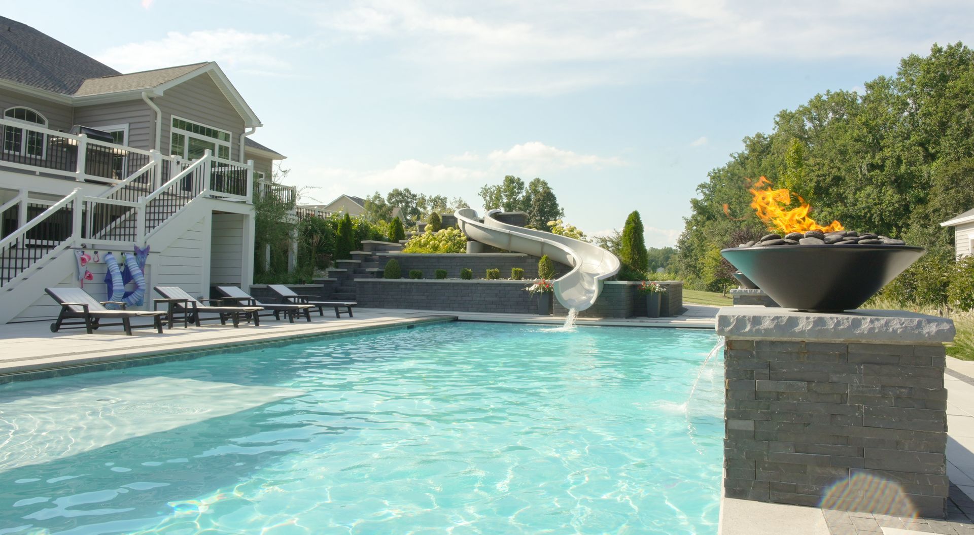 A luxurious backyard pool area with a fire pit, slide, and seating next to a large house under a bright sky.