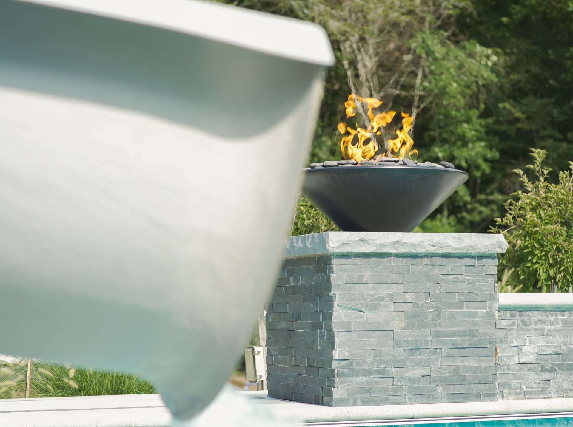 A fire pit with flames sits on a stone pillar near a pool, with the side of a white object in the foreground.