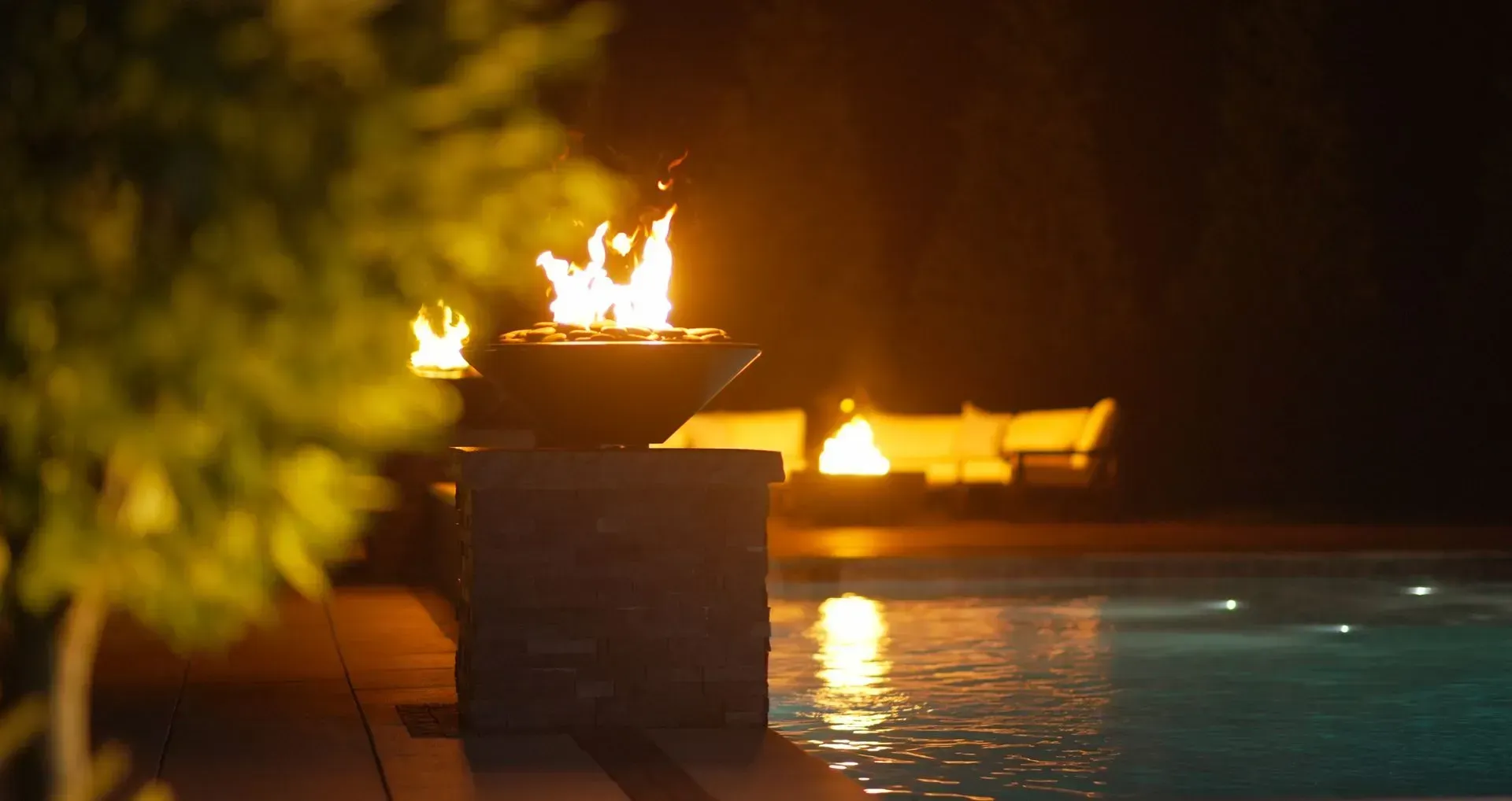 A fire pit with flames lit near a pool at night