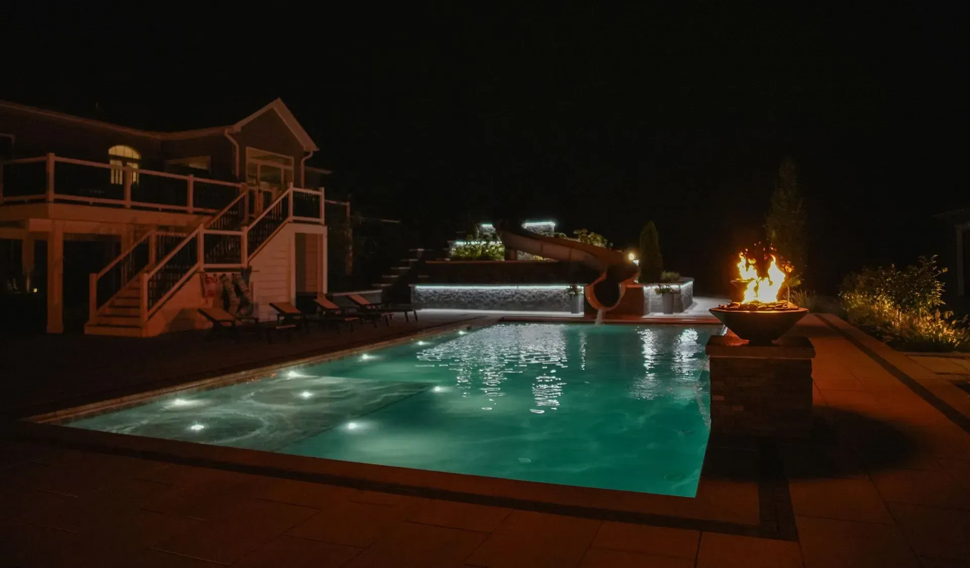 A pool with illuminated water, a lit fire pit, and a house with a deck