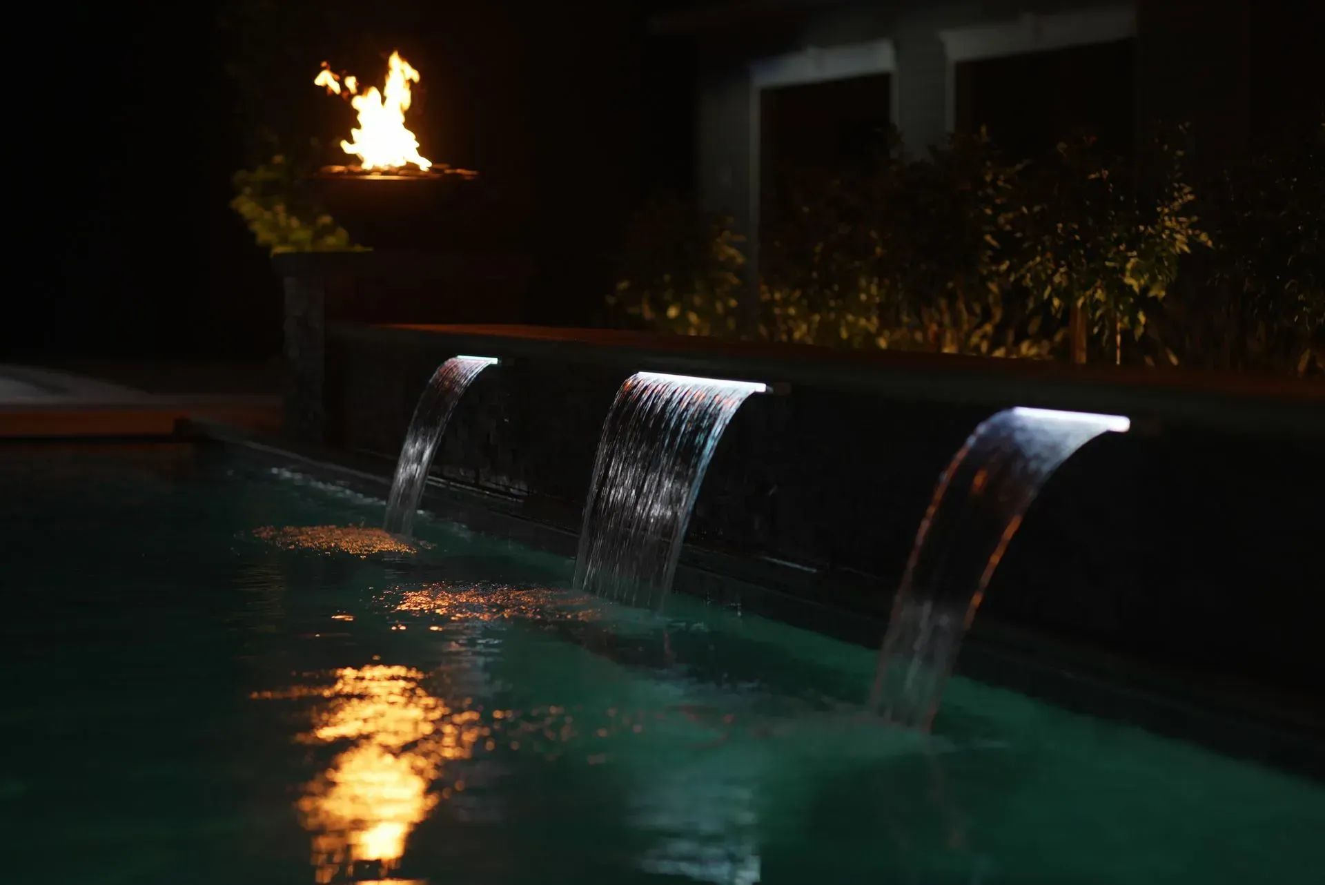 Pool with illuminated waterfalls and a fire feature at night