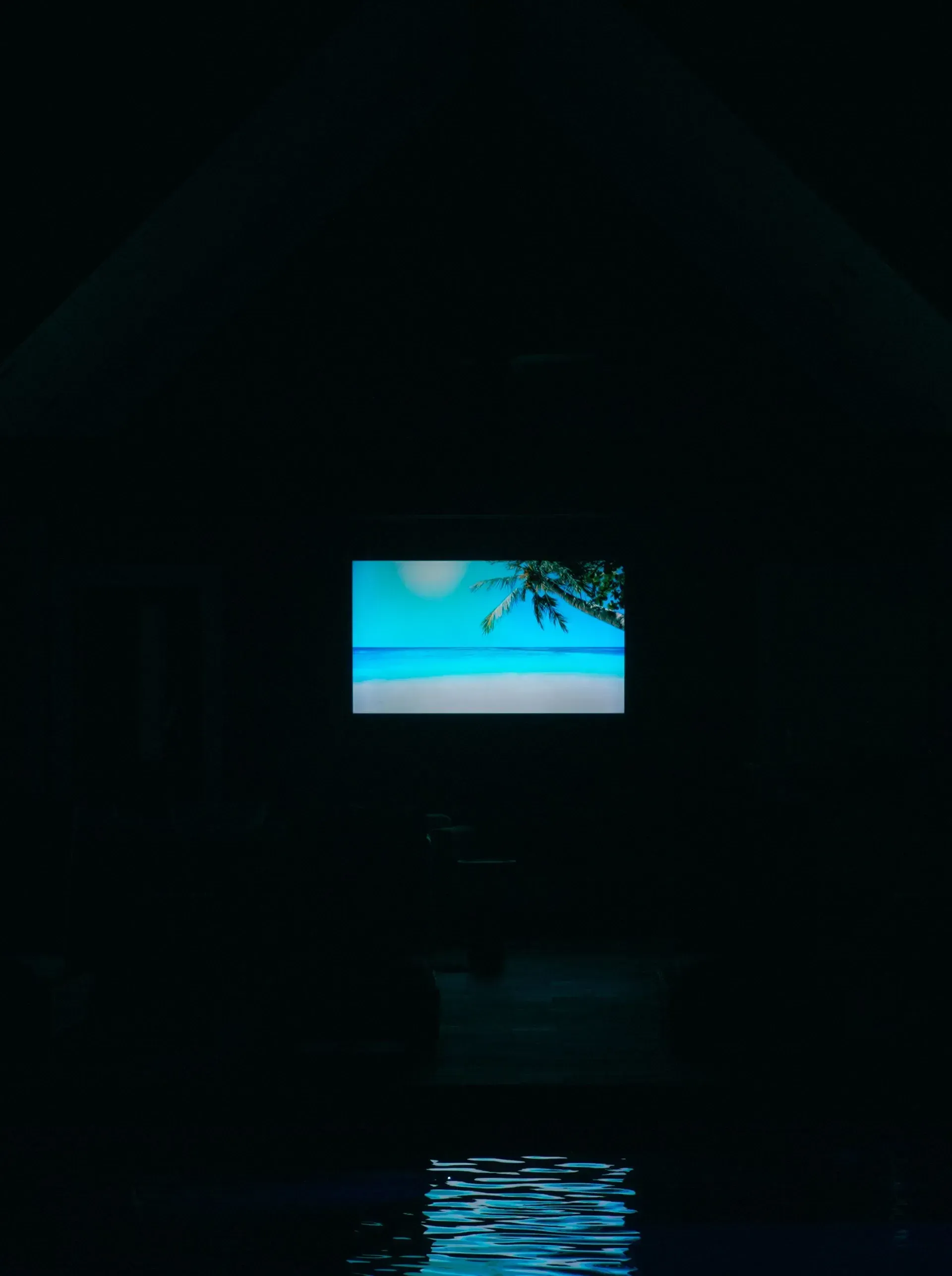 Dark room with a lit screen showing a beach scene, reflected in water below