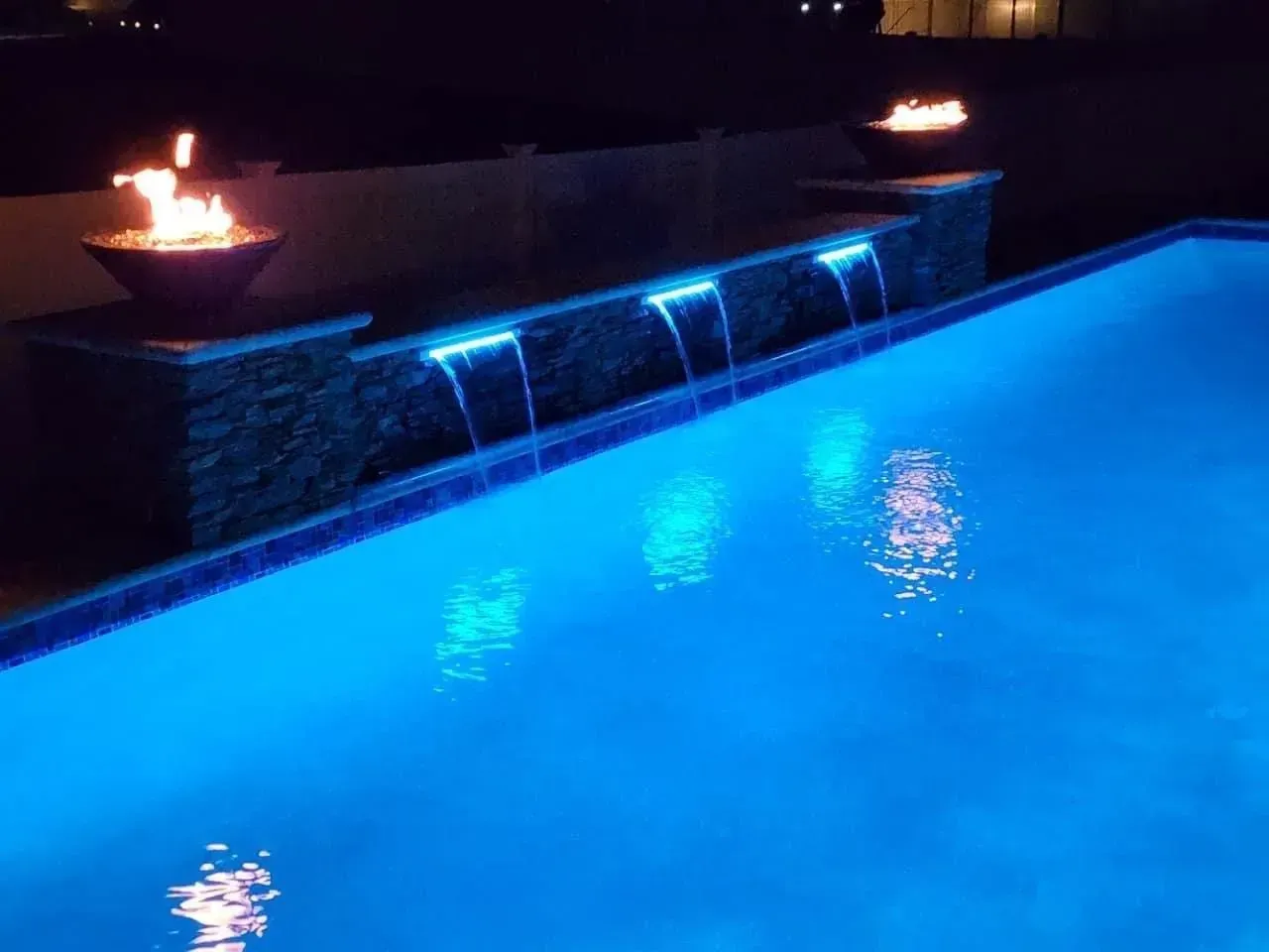 a swimming pool is lit up at night with a fire pit in the background