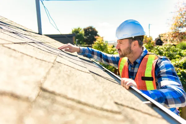 Worker in hard hat and safety vest inspecting a sunlit rooftop with a tool