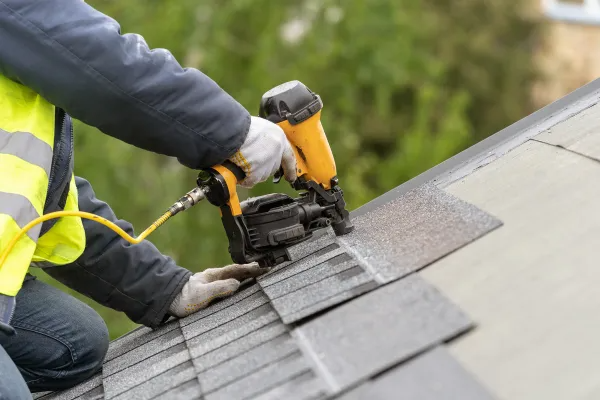 Worker using a yellow roofing nail gun to install shingles on a gray roof