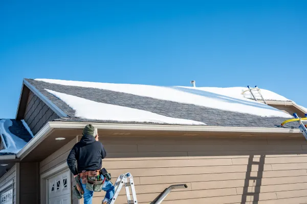 Person on a ladder cleaning snow from a house roof under a clear blue sky