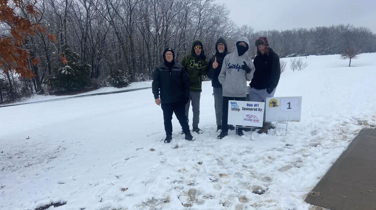 Four people posing in a snowy field beside a sign and marker on an overcast winter day
