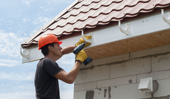 Worker in red cap repairs roof gutter with a drill under a tiled roof outdoors