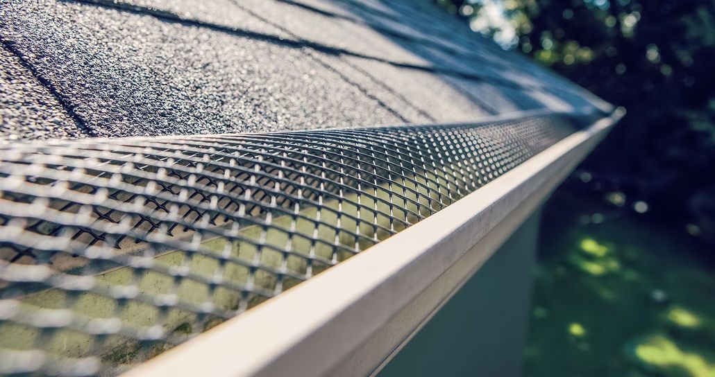 Close-up of a metal roof edge with gutter and shingles, viewed diagonally against blurred greenery below