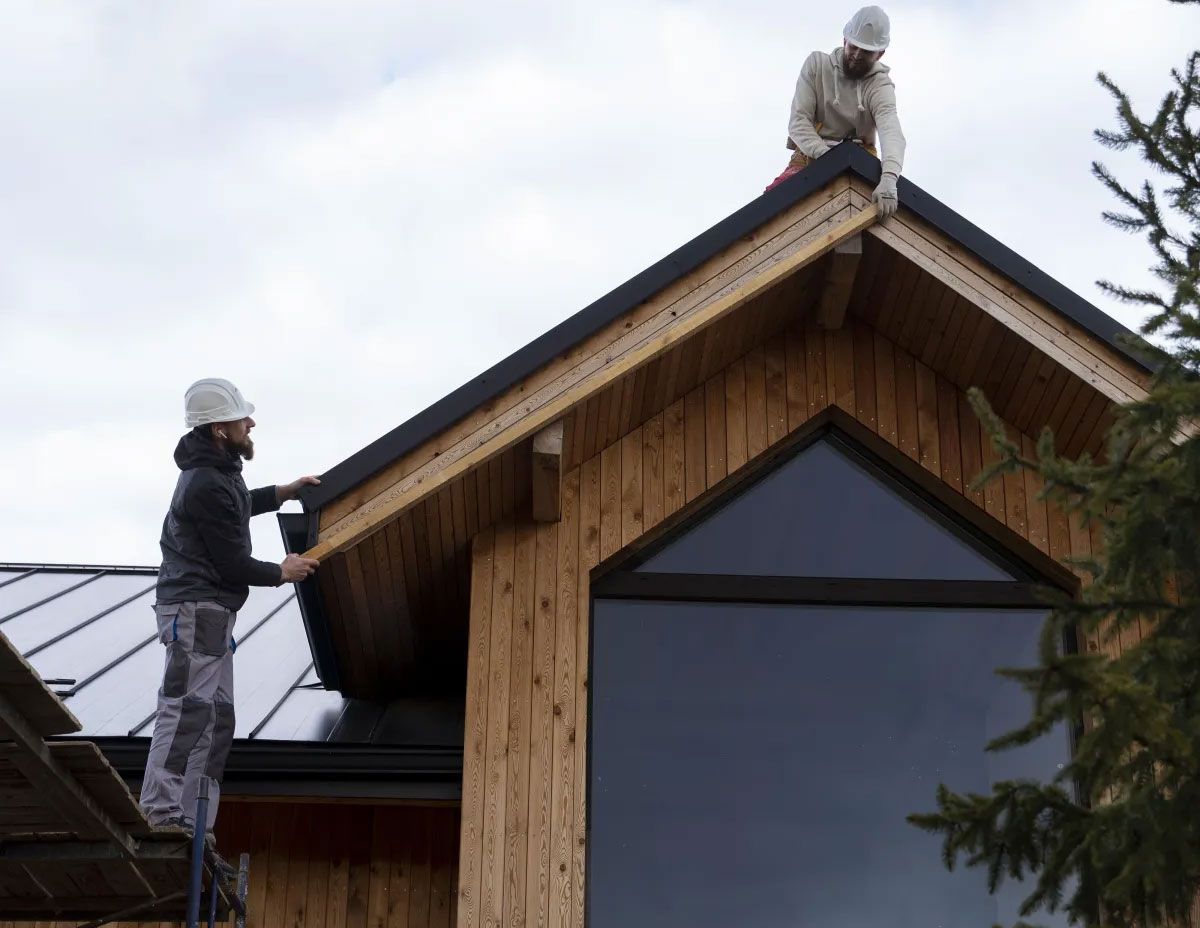 Two workers installing roofing on a wooden house, one on a ladder and one on the roof peak.