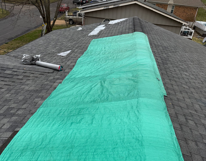 Green tarp covering a roof under repair, with tools and shingles visible on the gray rooftop.