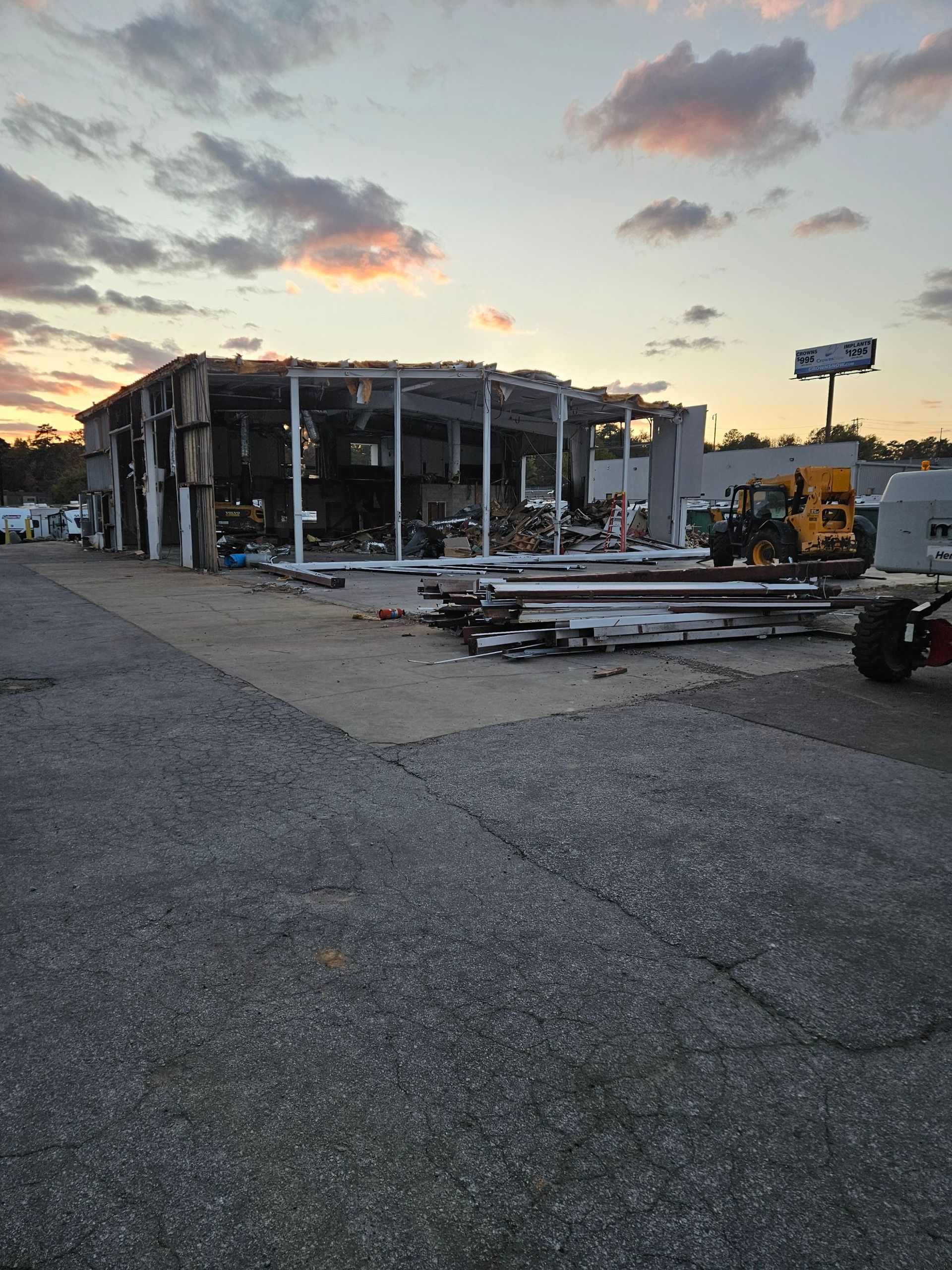 Damaged building at dusk with debris, exposed frame, and a yellow excavator in a gravel lot.