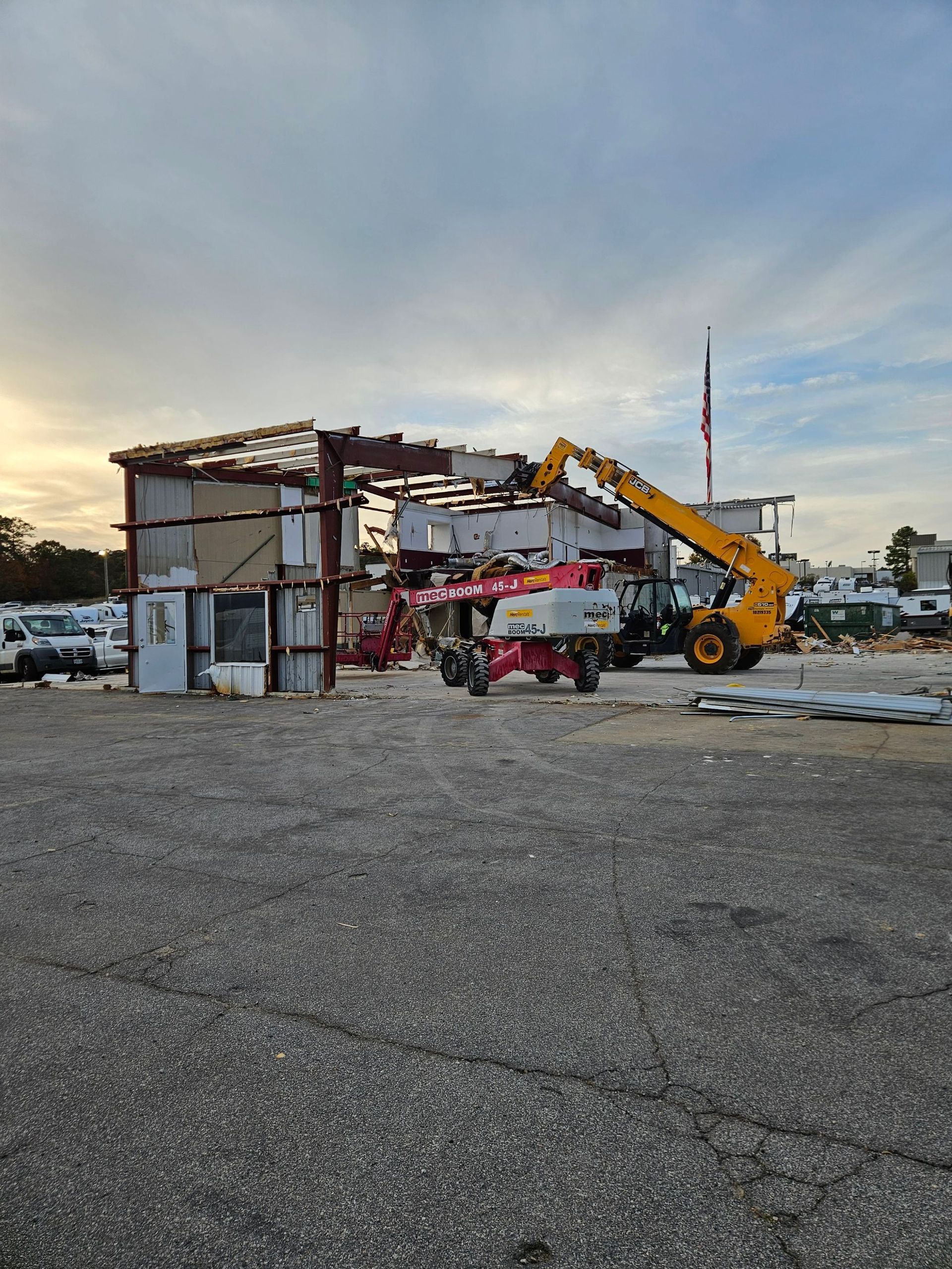 Demolition site with a partially dismantled building, yellow excavator, and debris on a gravel lot at sunset