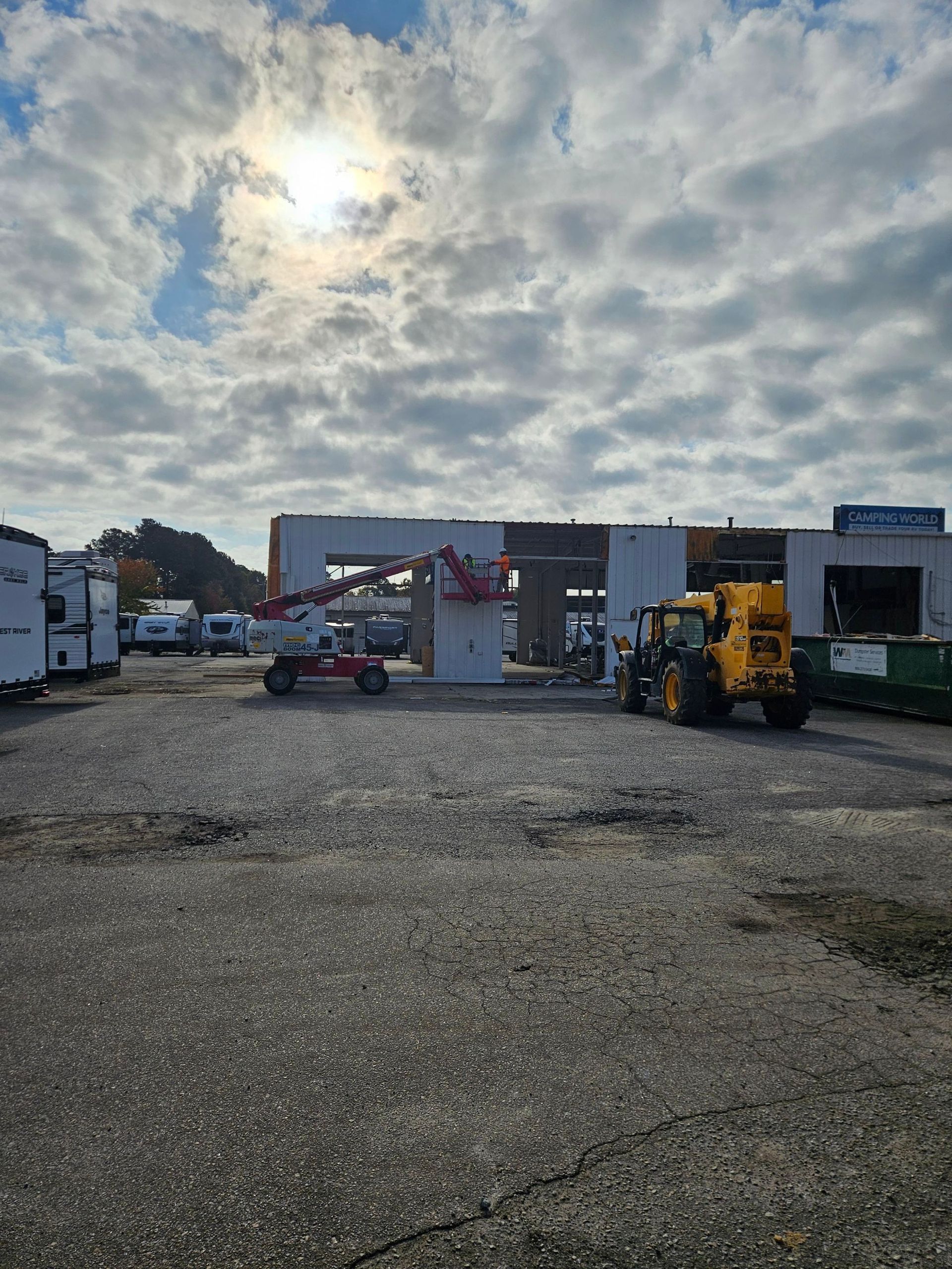 Gravel lot with parked trucks and equipment under a cloudy sky, with a small building in the background