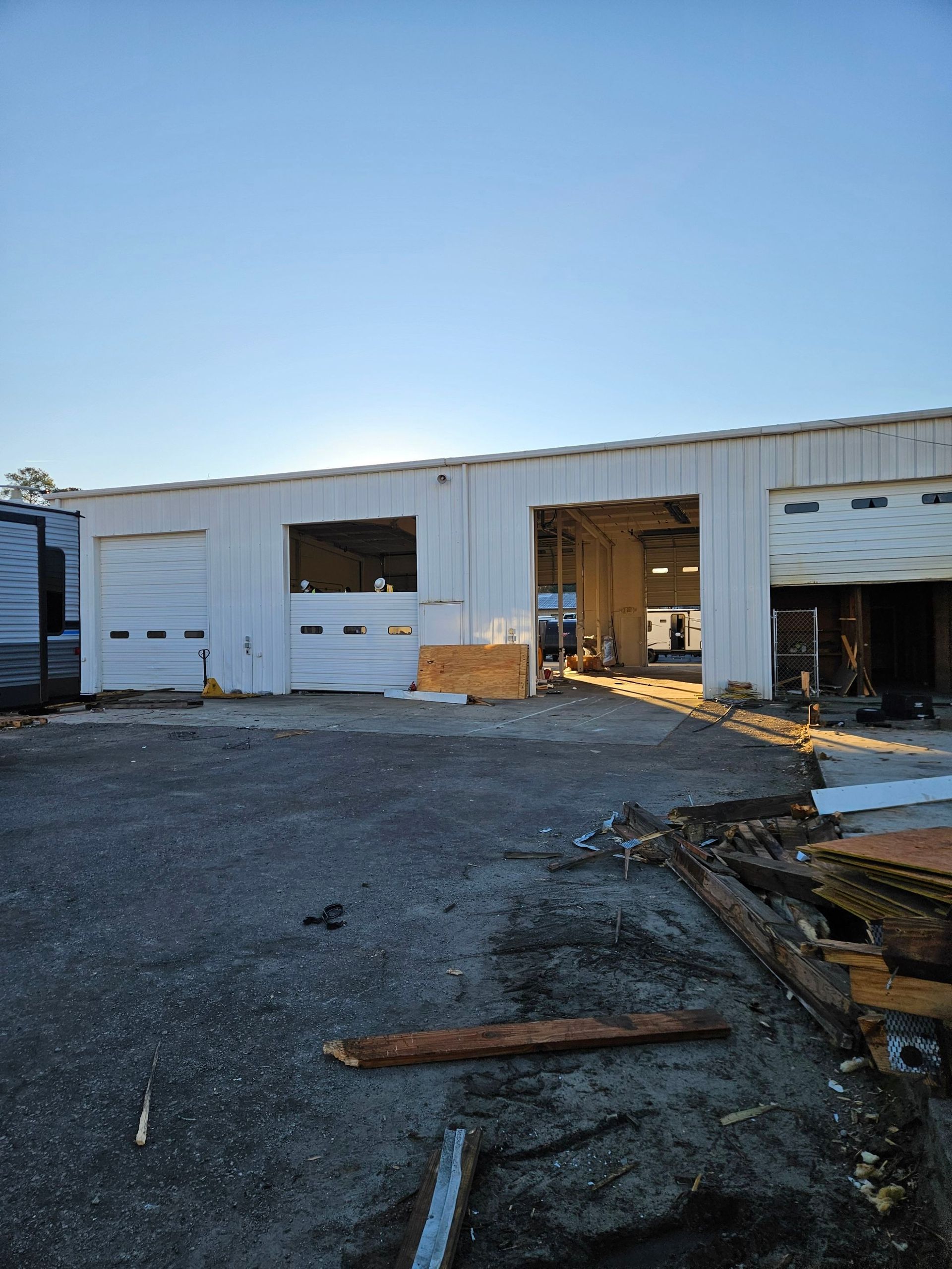 Damaged white industrial building with open bays and debris on a gravel lot under a clear sky