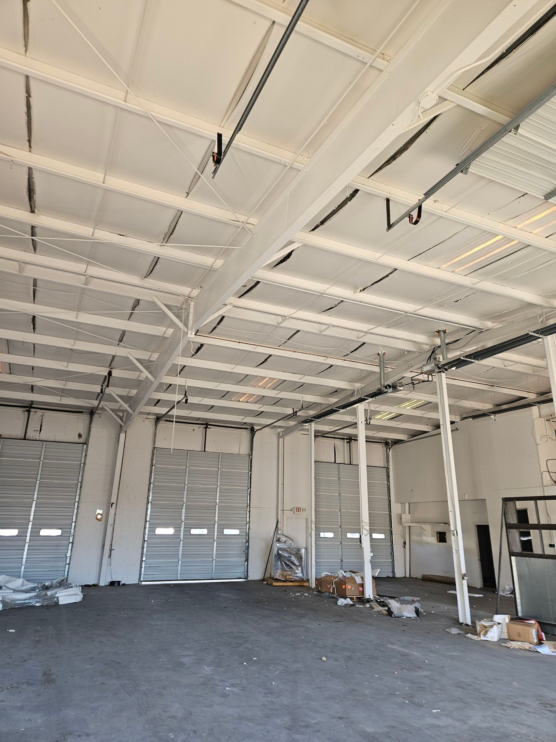 Empty industrial warehouse interior with white ceiling beams, roll-up doors, and scattered boxes on a concrete floor