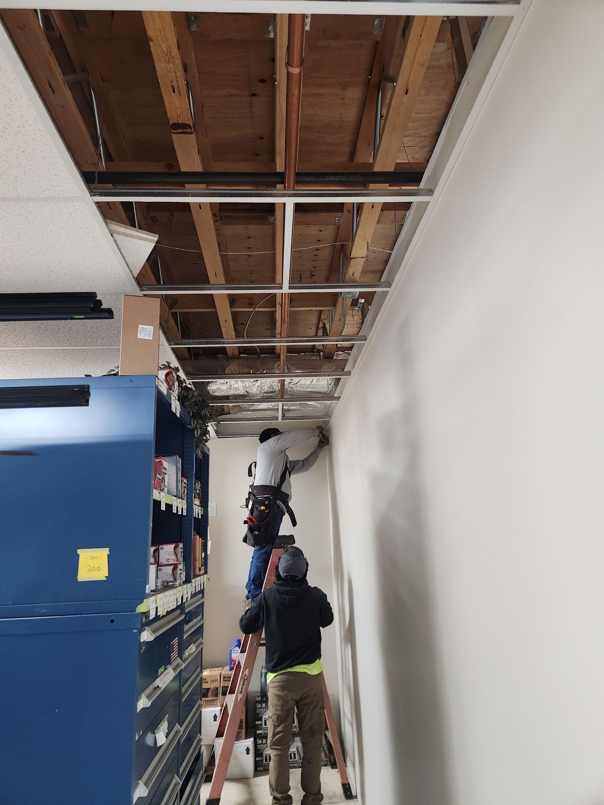 Workers on ladders repairing a ceiling with exposed beams in a hallway beside blue equipment.