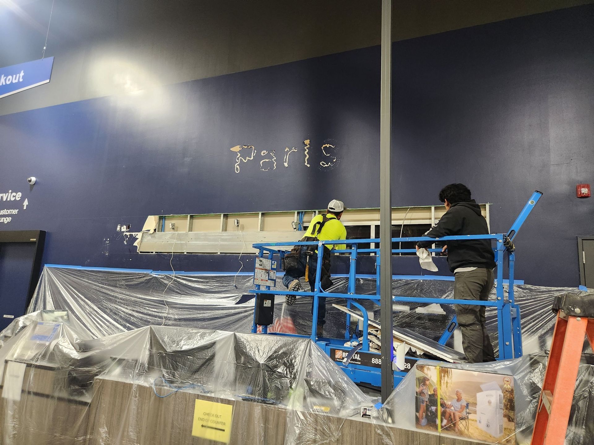 Workers on blue lifts repairing a wall in a large indoor space, with plastic sheeting below.