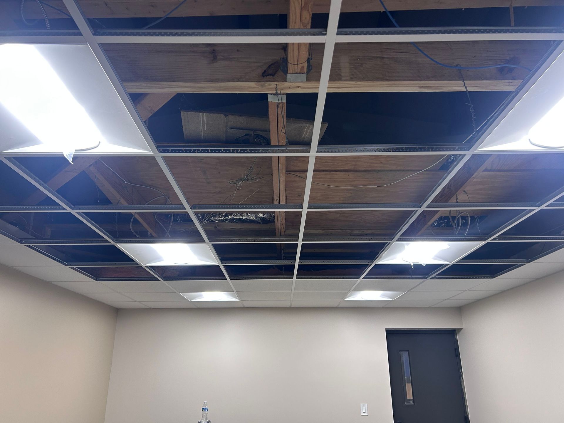Partially missing drop ceiling tiles in a room, exposing wiring and ductwork above fluorescent lights