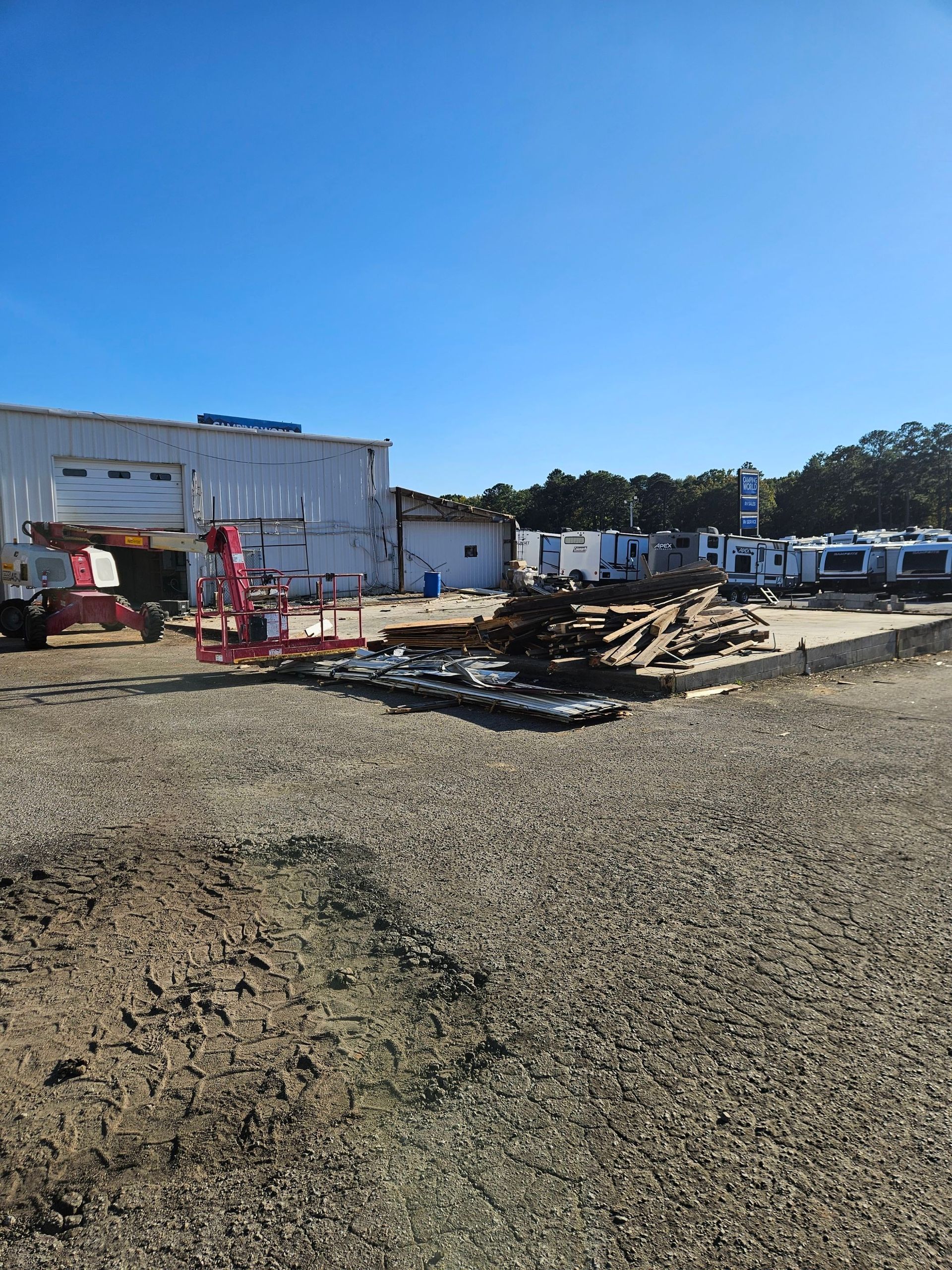 Construction site with gravel, debris pile, red machinery, and white buildings under a clear blue sky